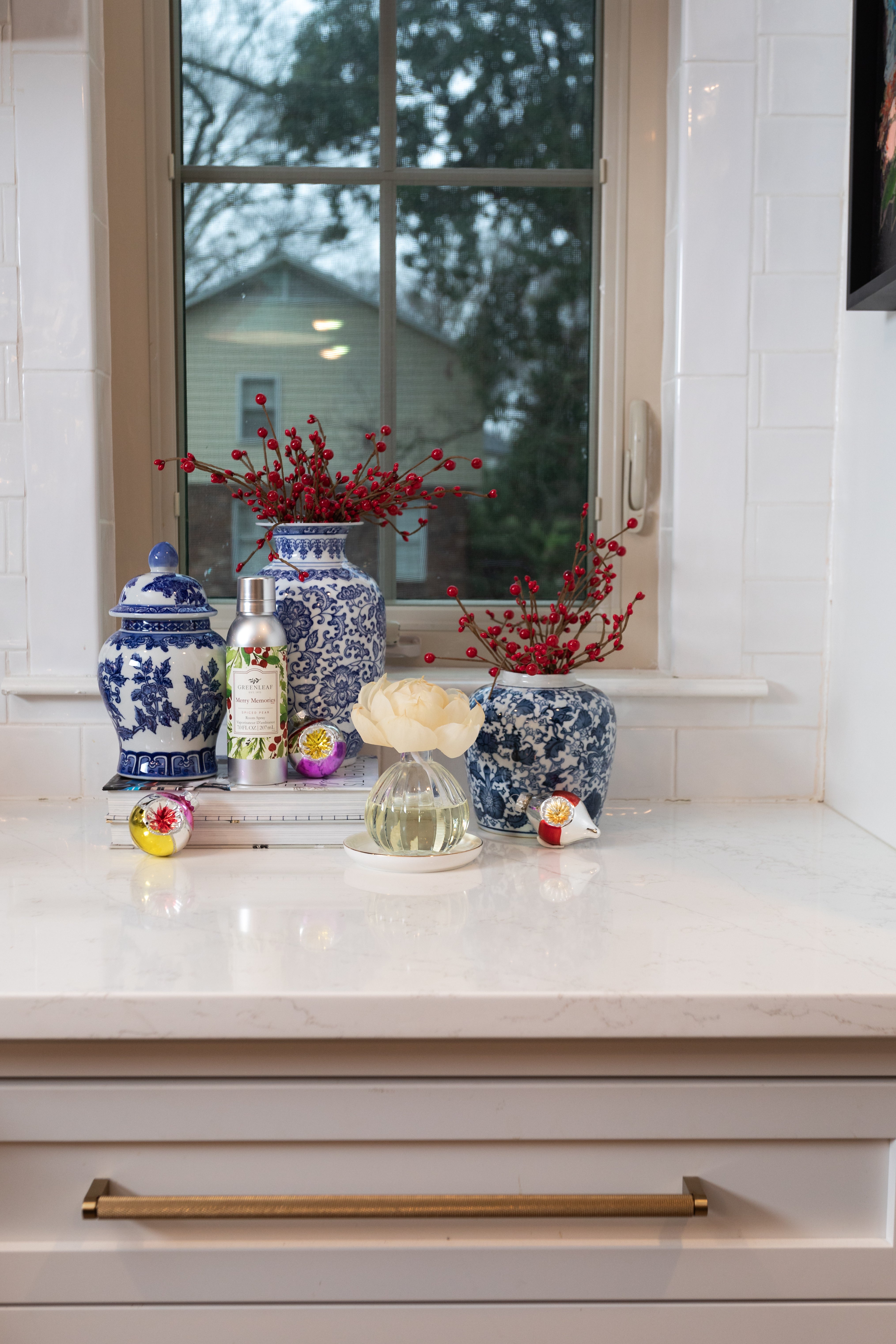 A kitchen counter with blue and white vases, red berries, a white flower in a glass, hand soap, soaps, and stacked books is refreshed by Greenleaf Gifts Merry Memories Room Spray, with a window view of a house and trees outside.