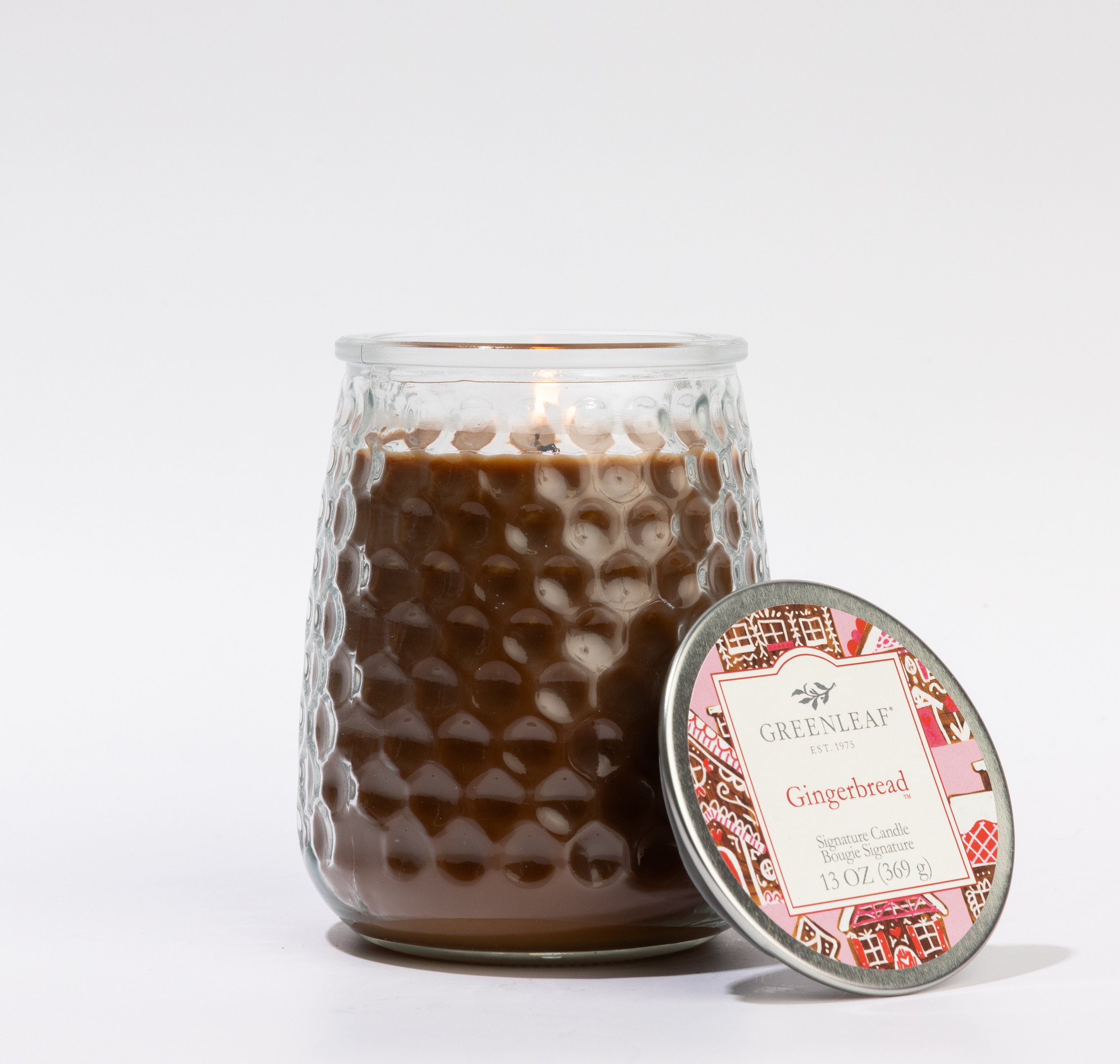 The Greenleaf Gifts Gingerbread Signature Candle is a 13 oz candle in a textured glass jar with a decorative-patterned lid, offering a cozy glow and inviting scent for 90-100 hours. Style: GLG915585S.