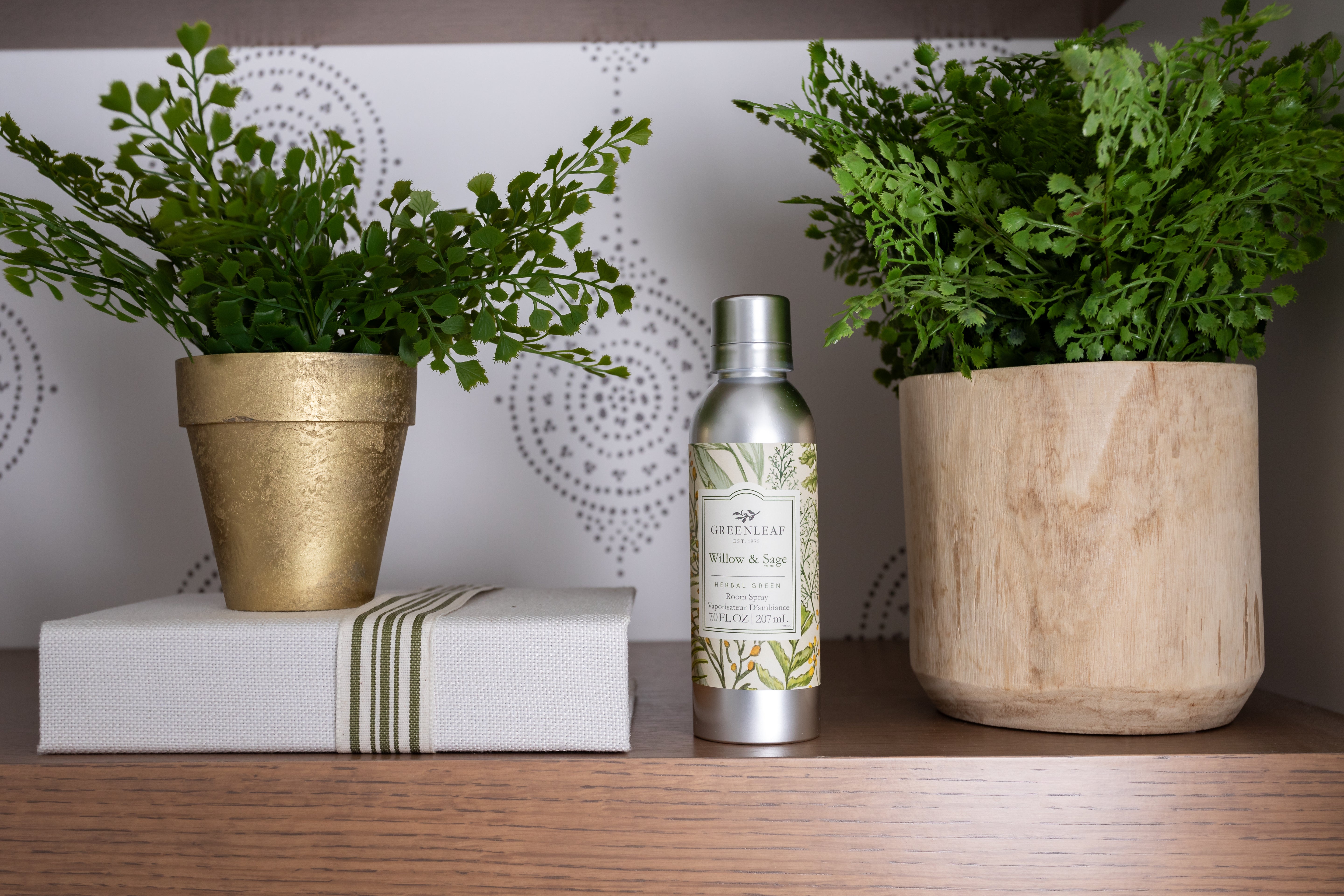 A Willow & Sage Room Spray by Greenleaf Gifts sits on a shelf between green potted plants—one in a gold pot on a white book, the other in wood—against a patterned wall, creating an elegant Greenleaf fragrance display.