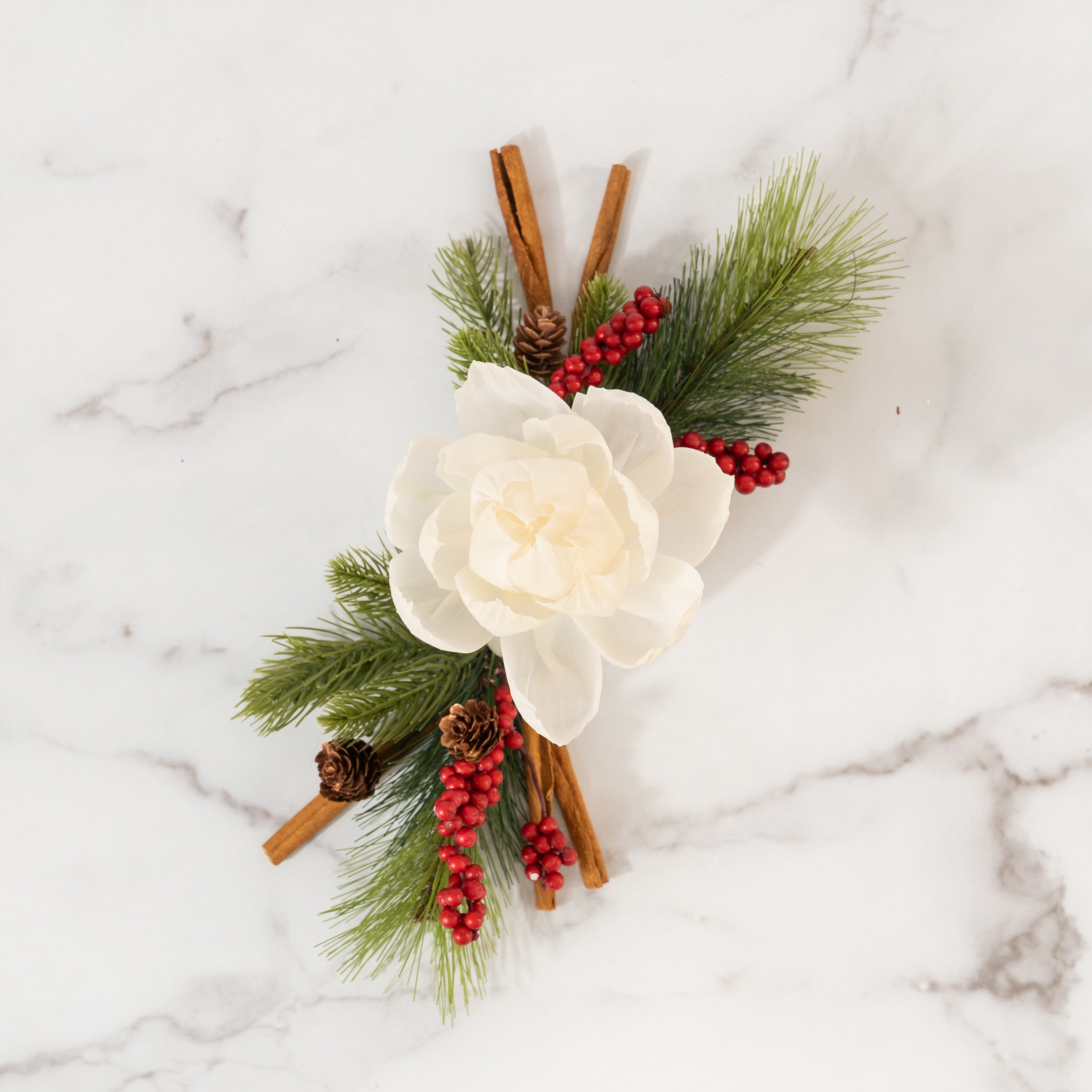 The Merry Memories Flower Diffuser by Greenleaf Gifts features a white flower with pine needles, red berries, pinecones, and cinnamon sticks on white marble. This festive arrangement offers up to 60 days of fragrance.