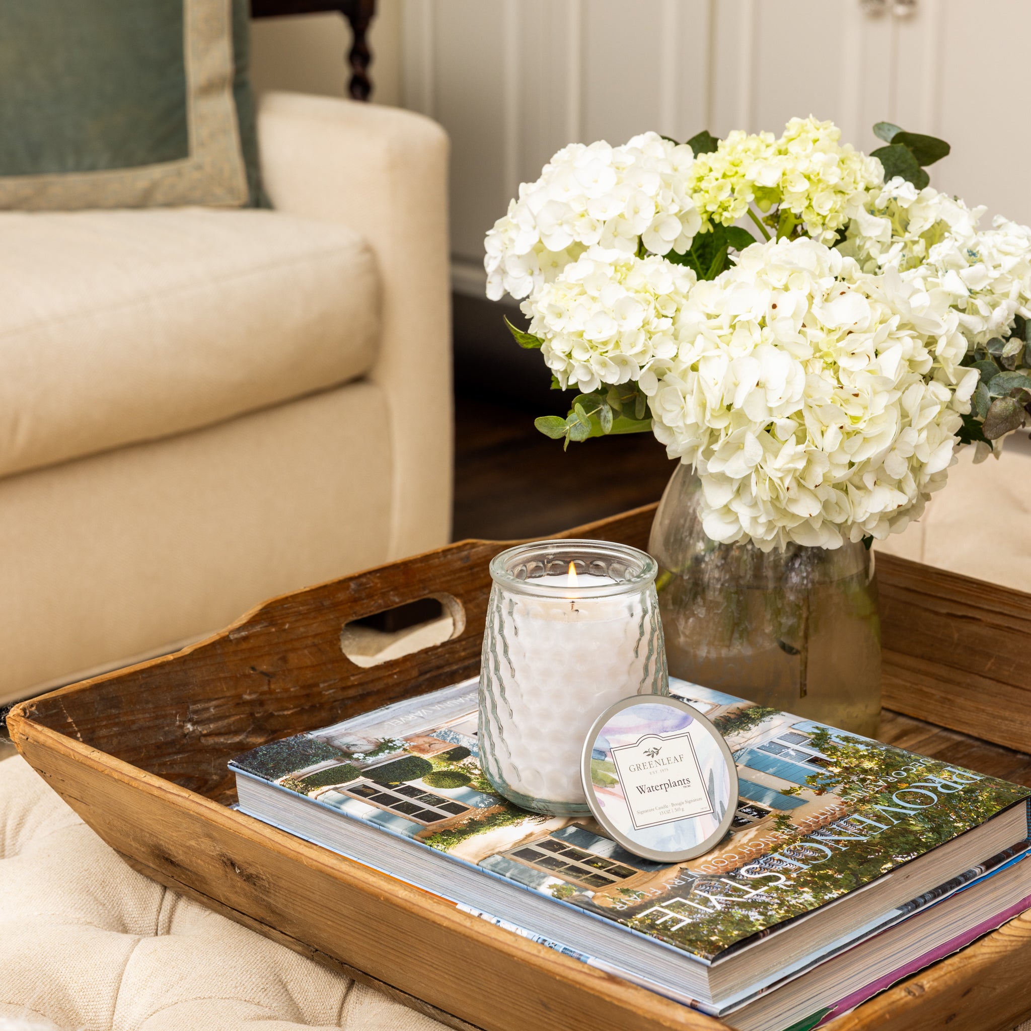 A wooden tray on an ottoman holds a vase of white hydrangeas, a Greenleaf Gifts Waterplants Signature Candle in a glass jar, and a large hardcover book, with a beige sofa and white cabinets in the background.