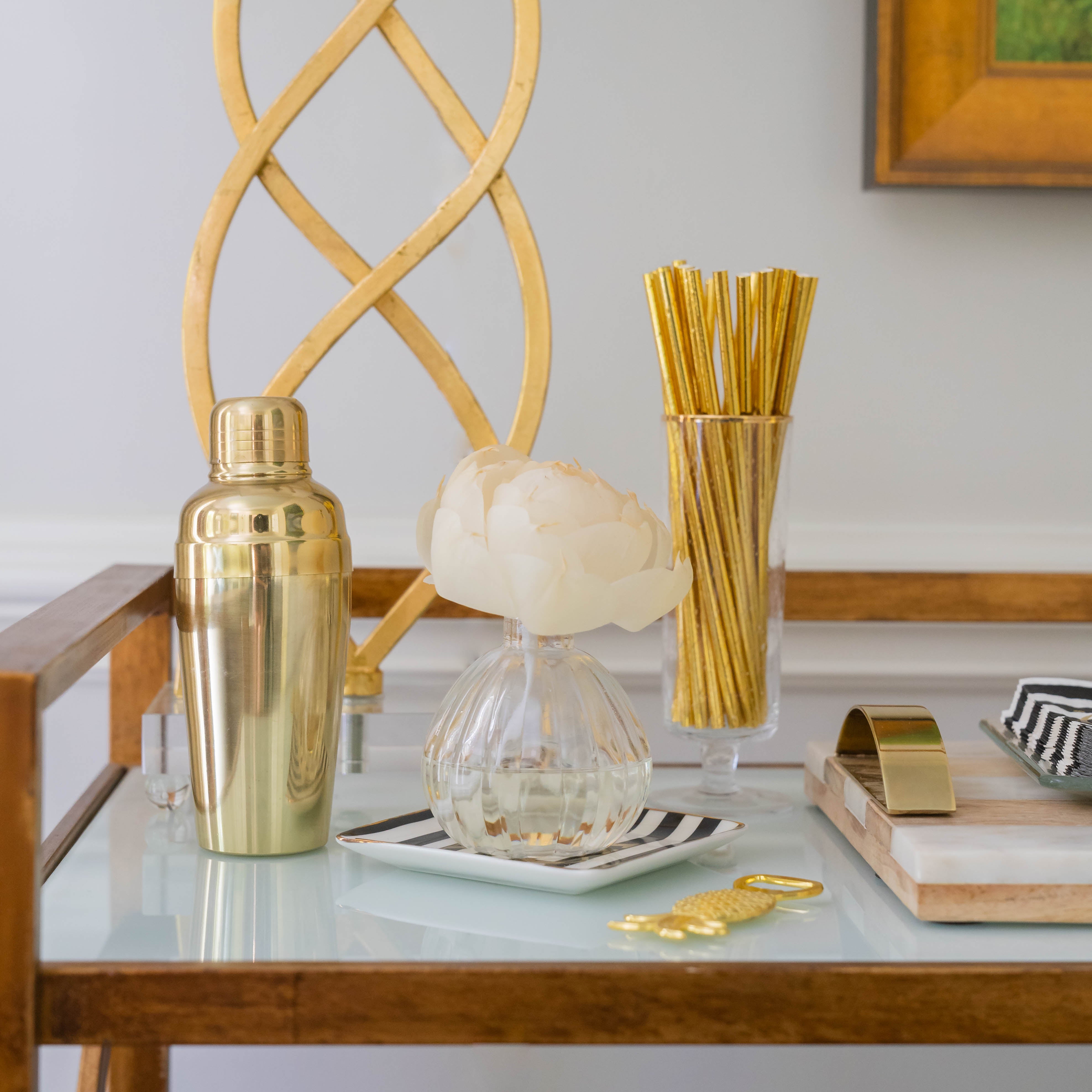 A stylish bar cart features a GLG966431S gold cocktail shaker, Greenleaf Gifts Flower Diffuser-Seaspray, gold straws in a clear holder, striped dish, marble tray, and gold bottle opener on a glass shelf—perfect for entertaining in style.