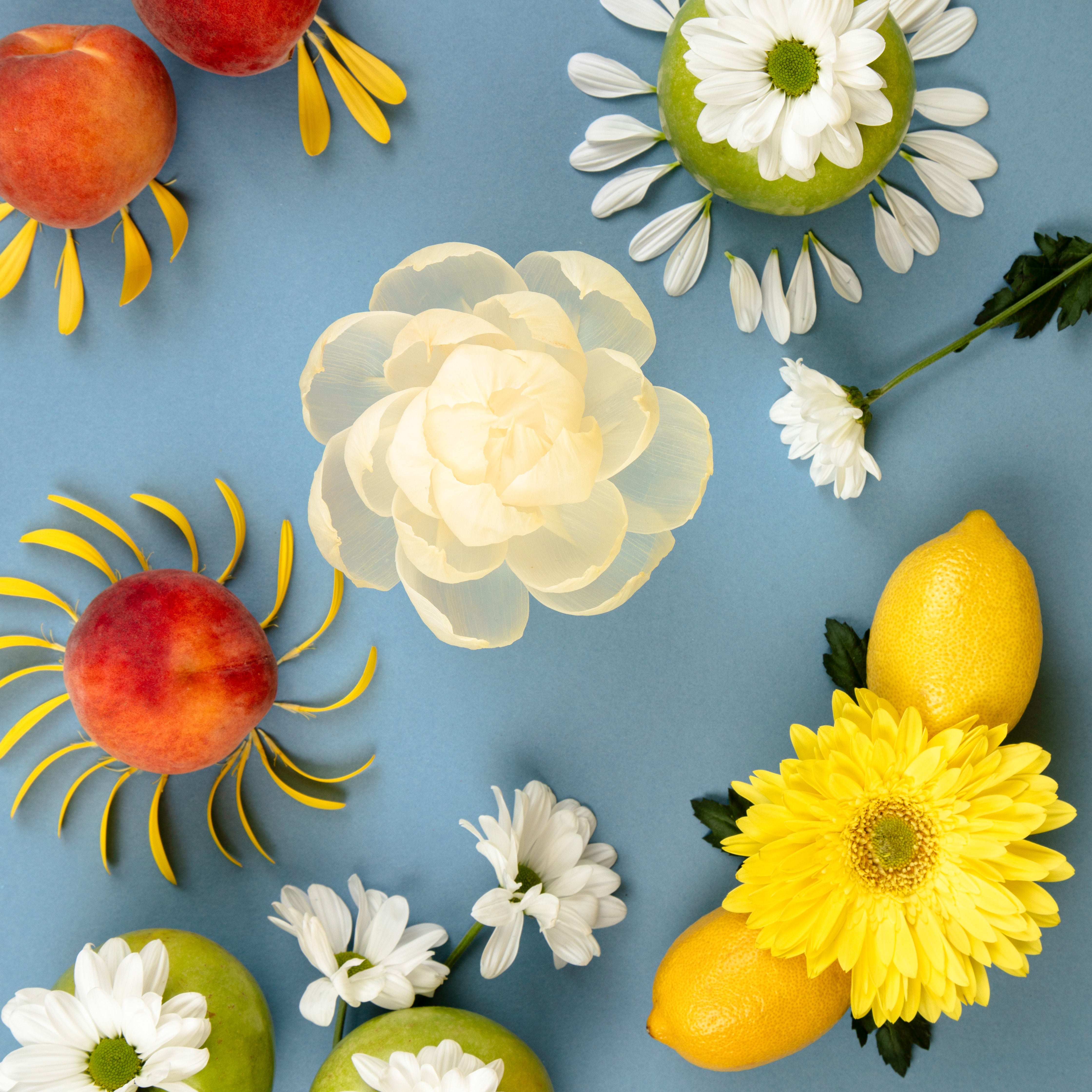 A flat lay of peaches, lemons, green apples, and white and yellow flowers on a blue background creates sun and floral patterns—inspiring decor for the Sunbeam Flower Diffuser by Greenleaf Gifts.