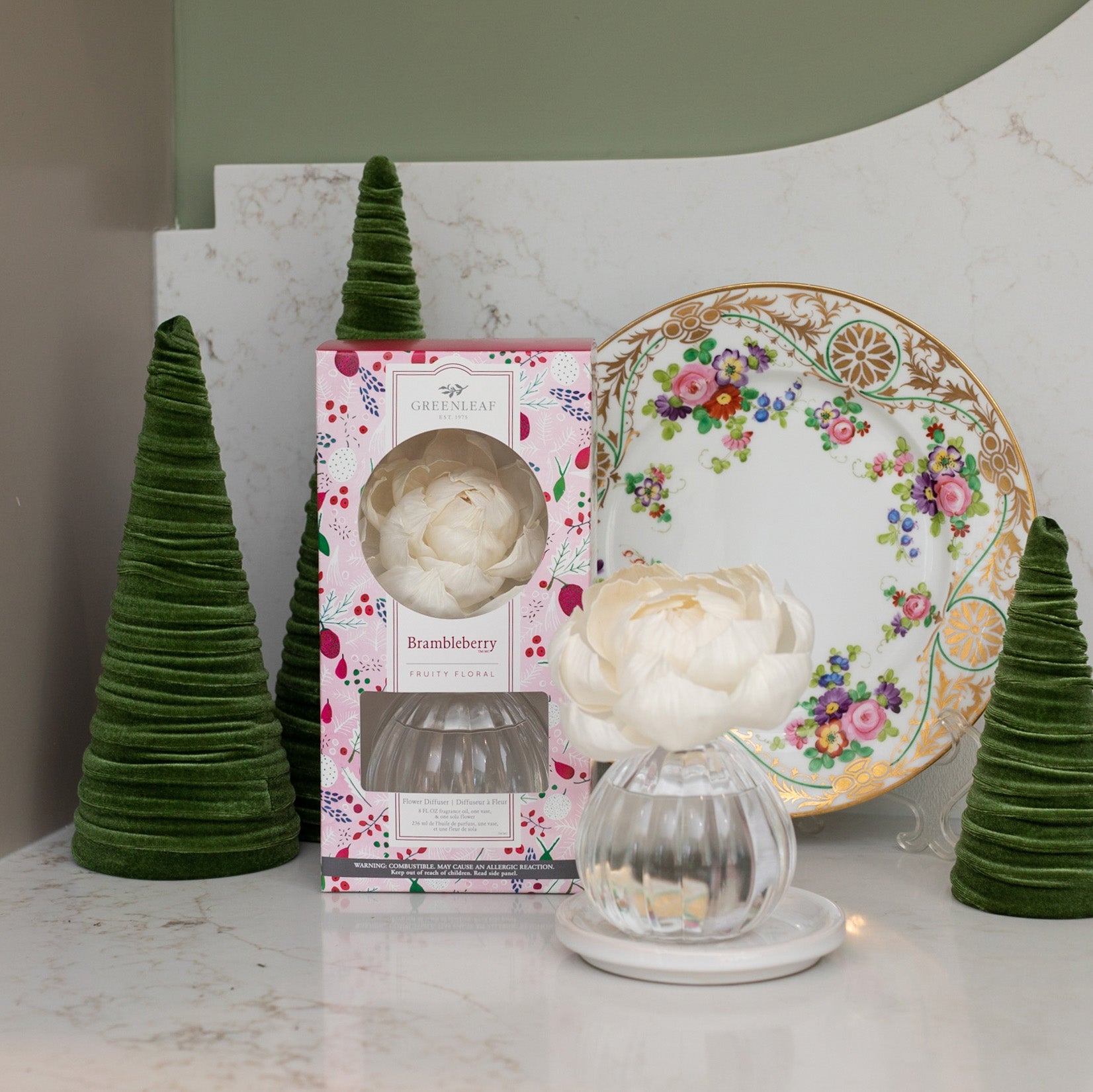 A decorative plate with a floral pattern stands behind three green velvet cone trees, the Greenleaf Gifts Brambleberry Flower Diffuser (8 oz, GLG966572S), and a diffuser jar with a white flower on marble. Enjoy up to 60 days of freshness.