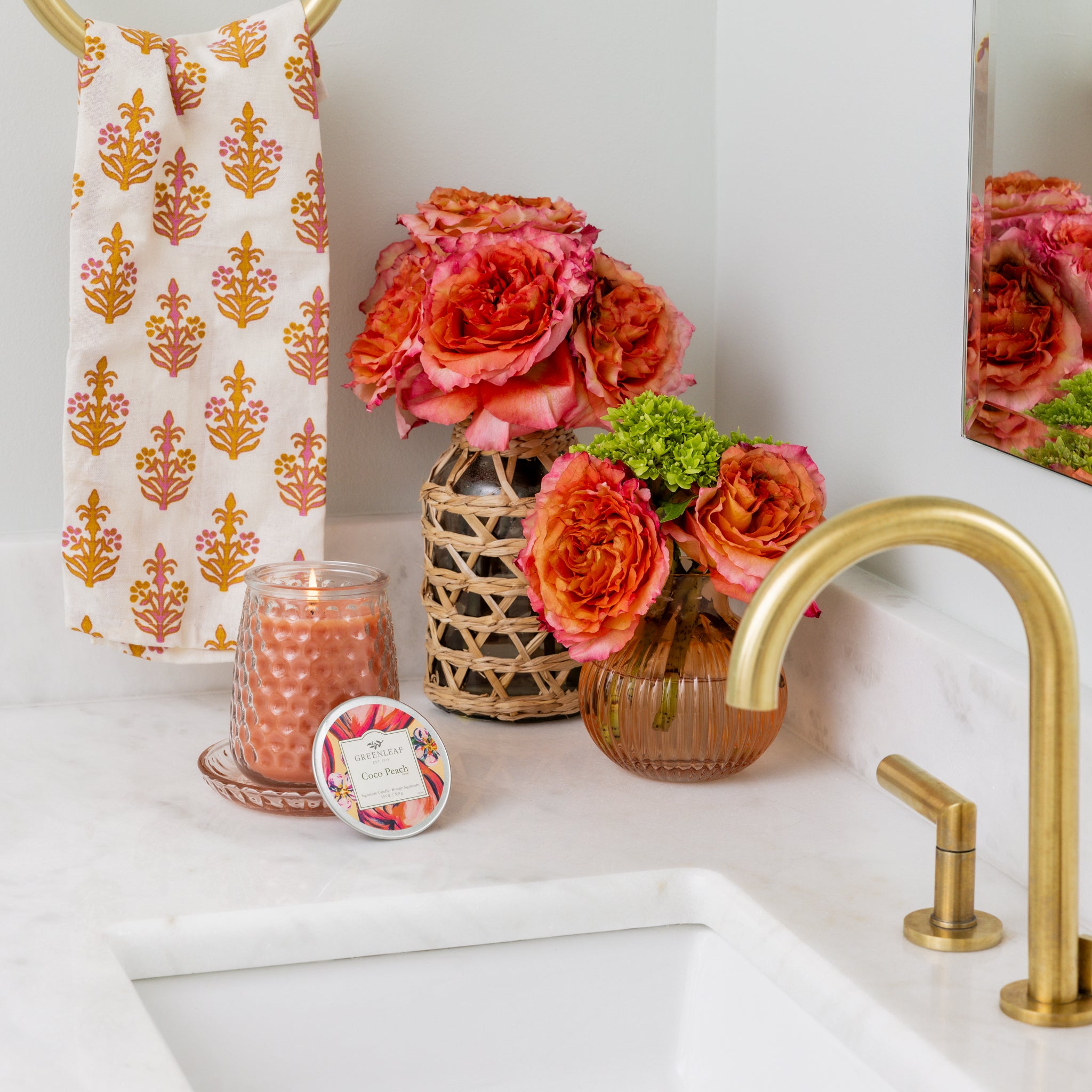 A bathroom countertop with a gold faucet, Greenleaf Gifts’ Coco Peach Signature Candle, a pink and orange floral arrangement in two vases, a small tin, and an orange and pink hand towel on a gold ring.