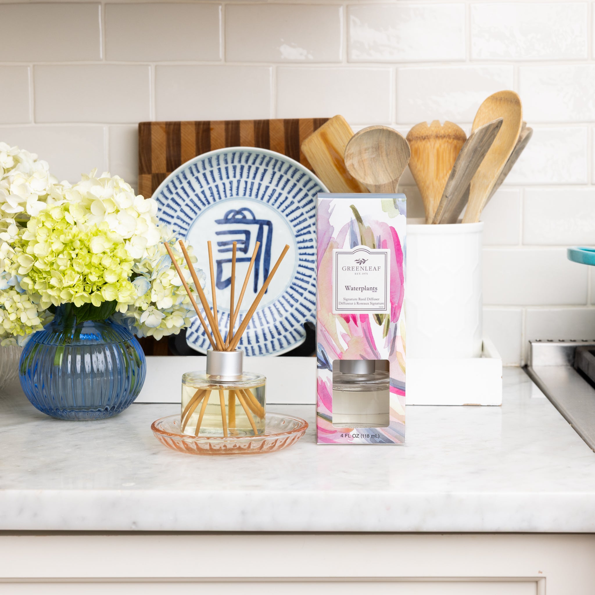 The Greenleaf Gifts Waterplants Diffuser Refill Oil, its box, a blue vase with white flowers, a blue and white plate, and a white utensil holder with wooden spoons sit on a marble kitchen counter against white tile.