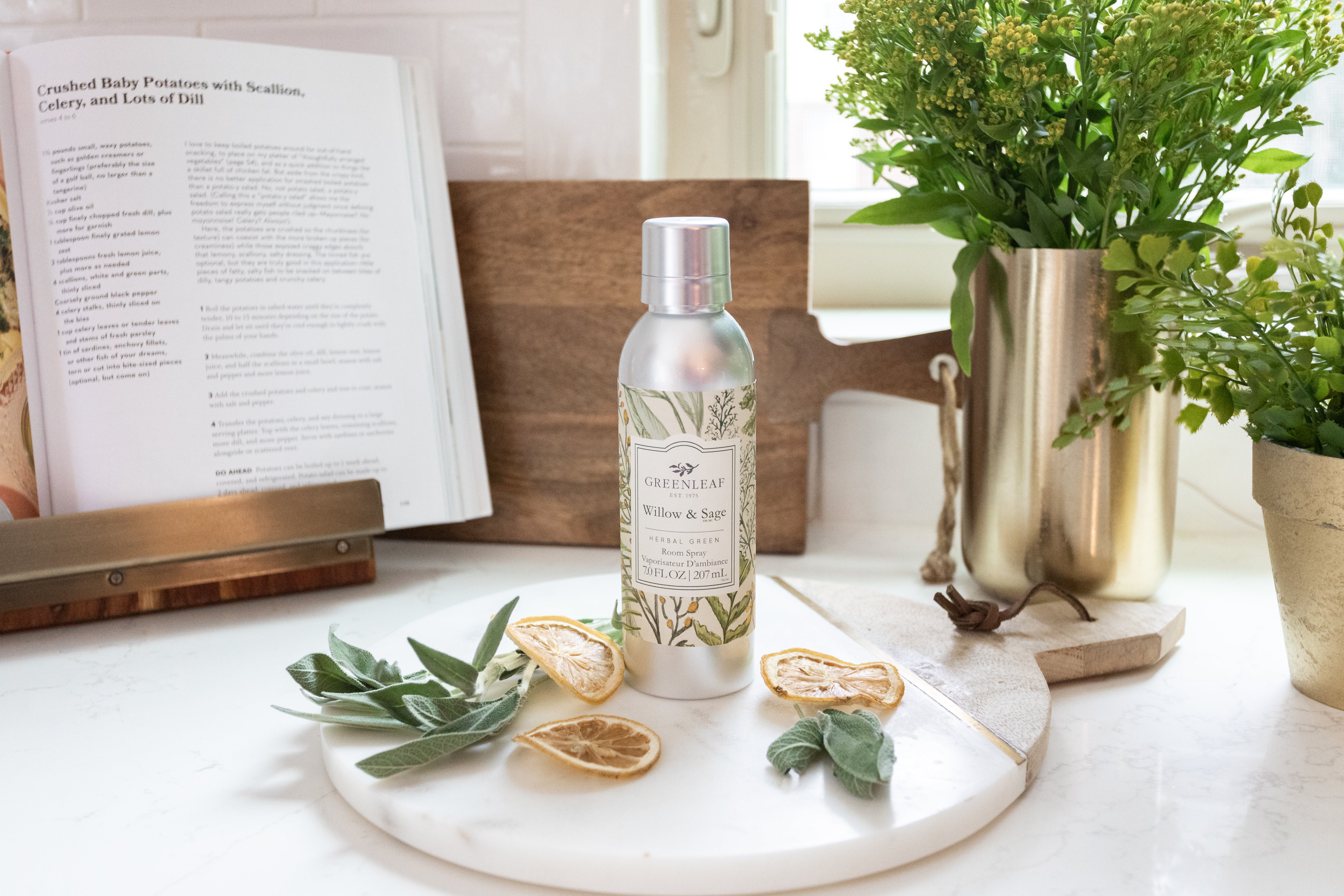 A silver Greenleaf Gifts Willow & Sage Room Spray rests on a white tray with dried citrus slices and sage leaves, set before a gold vase of greenery and an open cookbook on a kitchen counter.