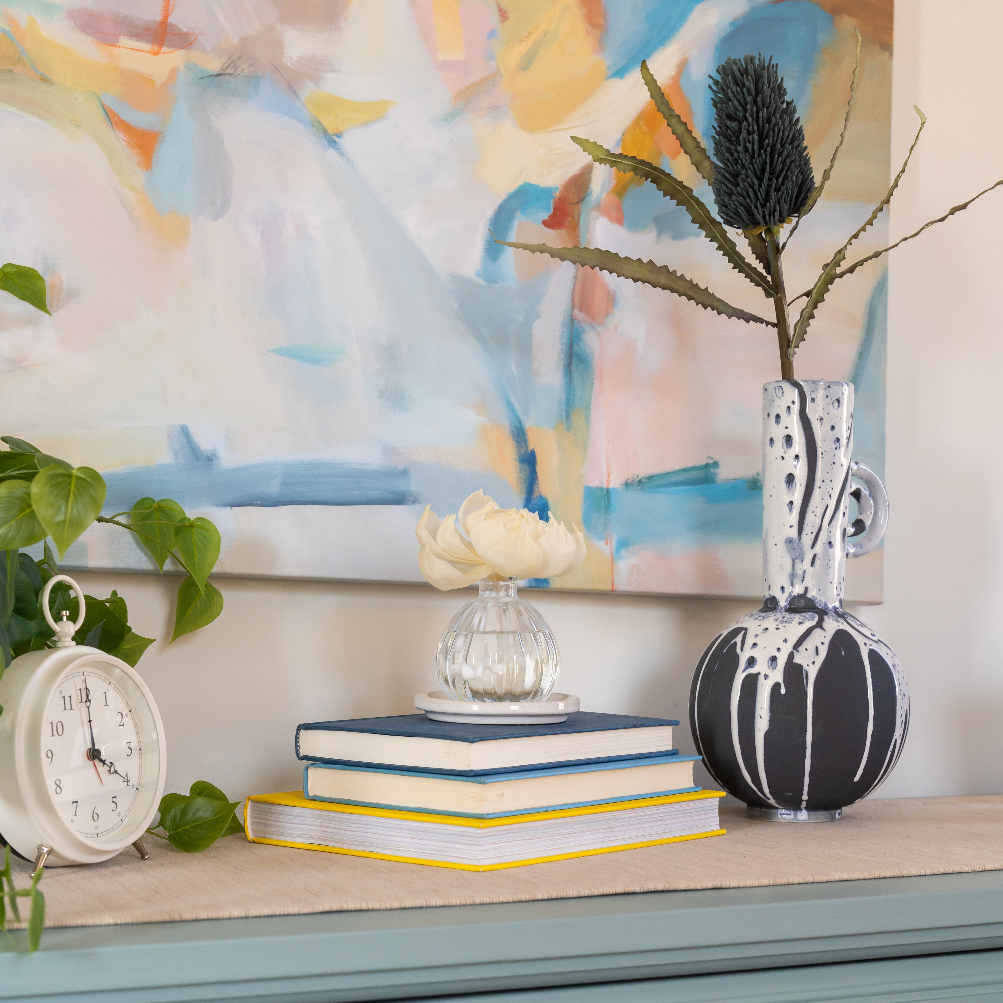 A tabletop displays a white clock, a small plant, books, and the Greenleaf Gifts Flower Diffuser-Classic Linen. A tall decorative vase with foliage is nearby, while an abstract painting hangs in the background.