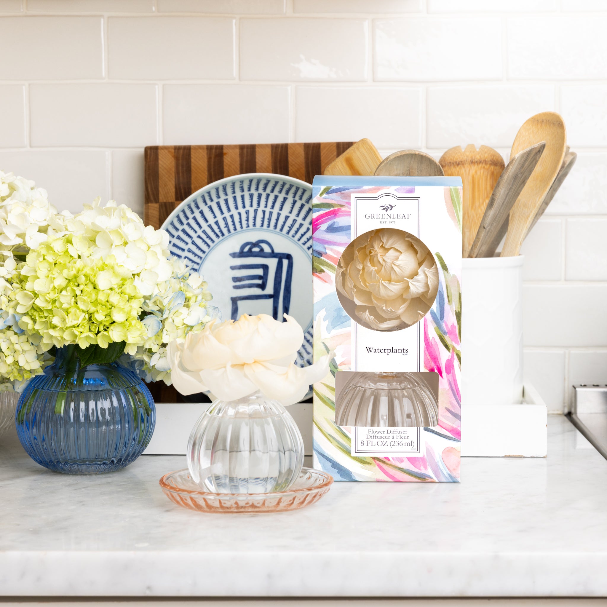 A kitchen counter displays a box labeled Greenleaf Gifts Waterplants Flower Diffuser next to a decorative vase with a sola paper flower, a blue and white plate, cutting boards, wooden utensils, and a blue vase of hydrangeas.