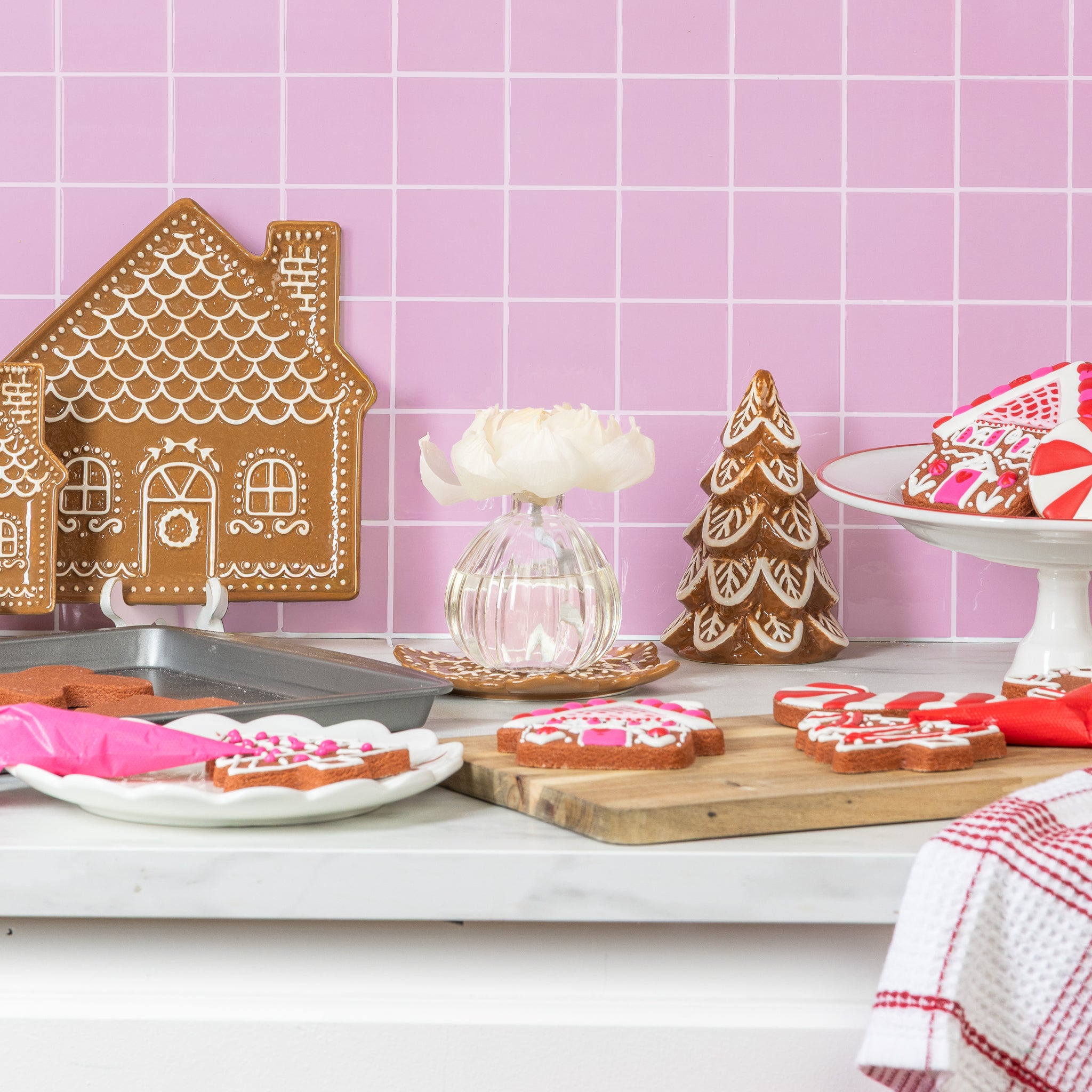 A kitchen counter with decorated gingerbread cookies, a house and tree, treat plate, icing bags, and a Greenleaf Gifts Gingerbread Flower Diffuser—sola paper flower in a glass vase—all set against a pink tiled wall.