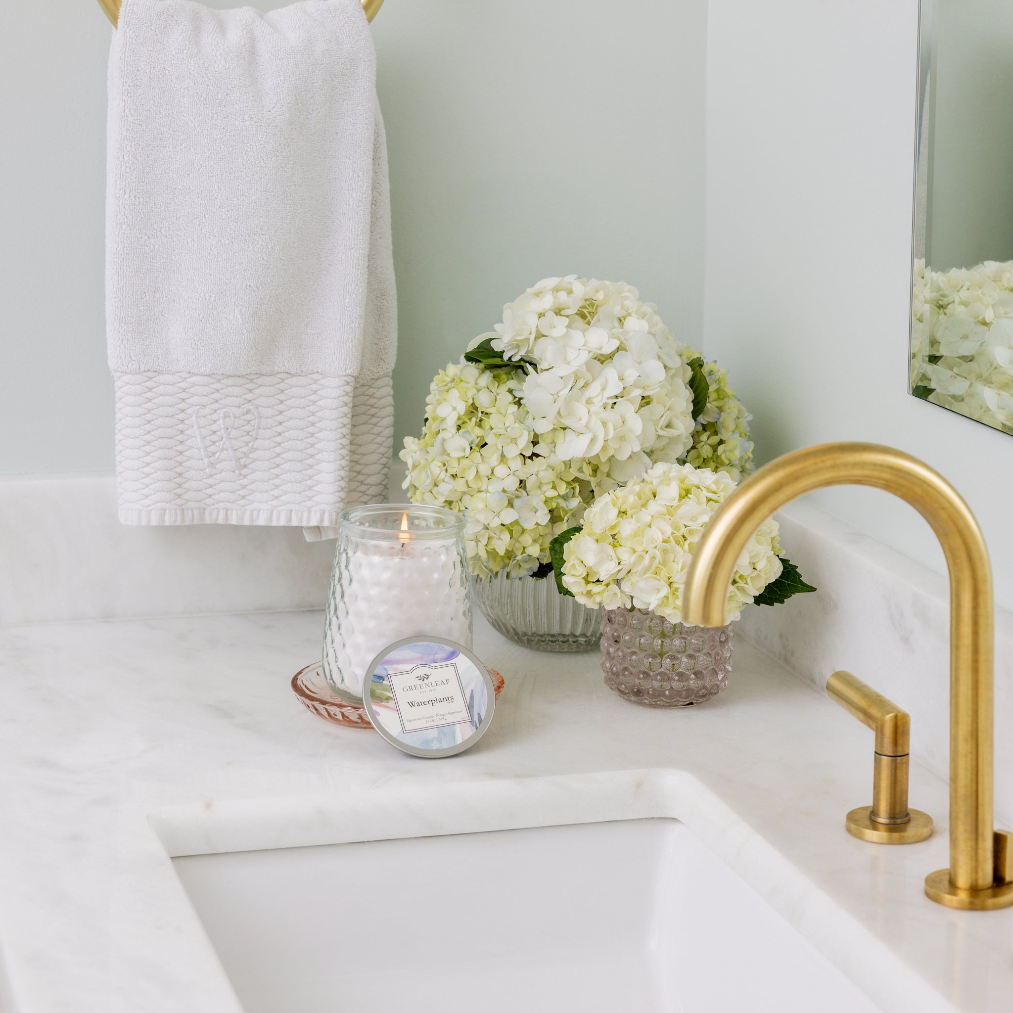 A bathroom countertop with a gold faucet, white sink, Greenleaf Gifts Waterplants Signature Candle, white hydrangeas in glass vases, and a white towel on a gold ring against a pale wall.