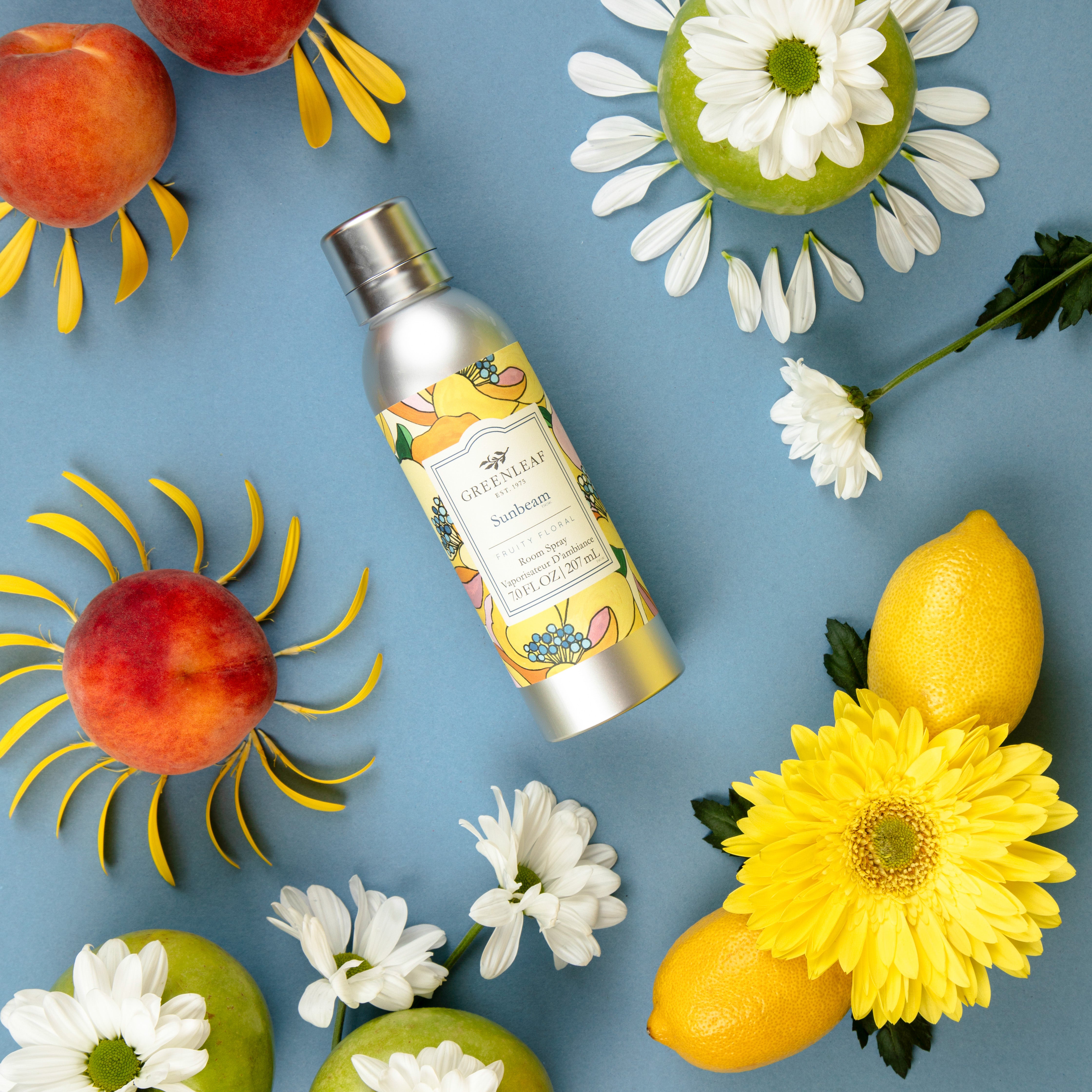 A bottle of Greenleaf Gifts Sunbeam Room Spray is displayed on a blue background with lemons, peaches, daisies, and green apples decorated with flower petals in sun and floral patterns.