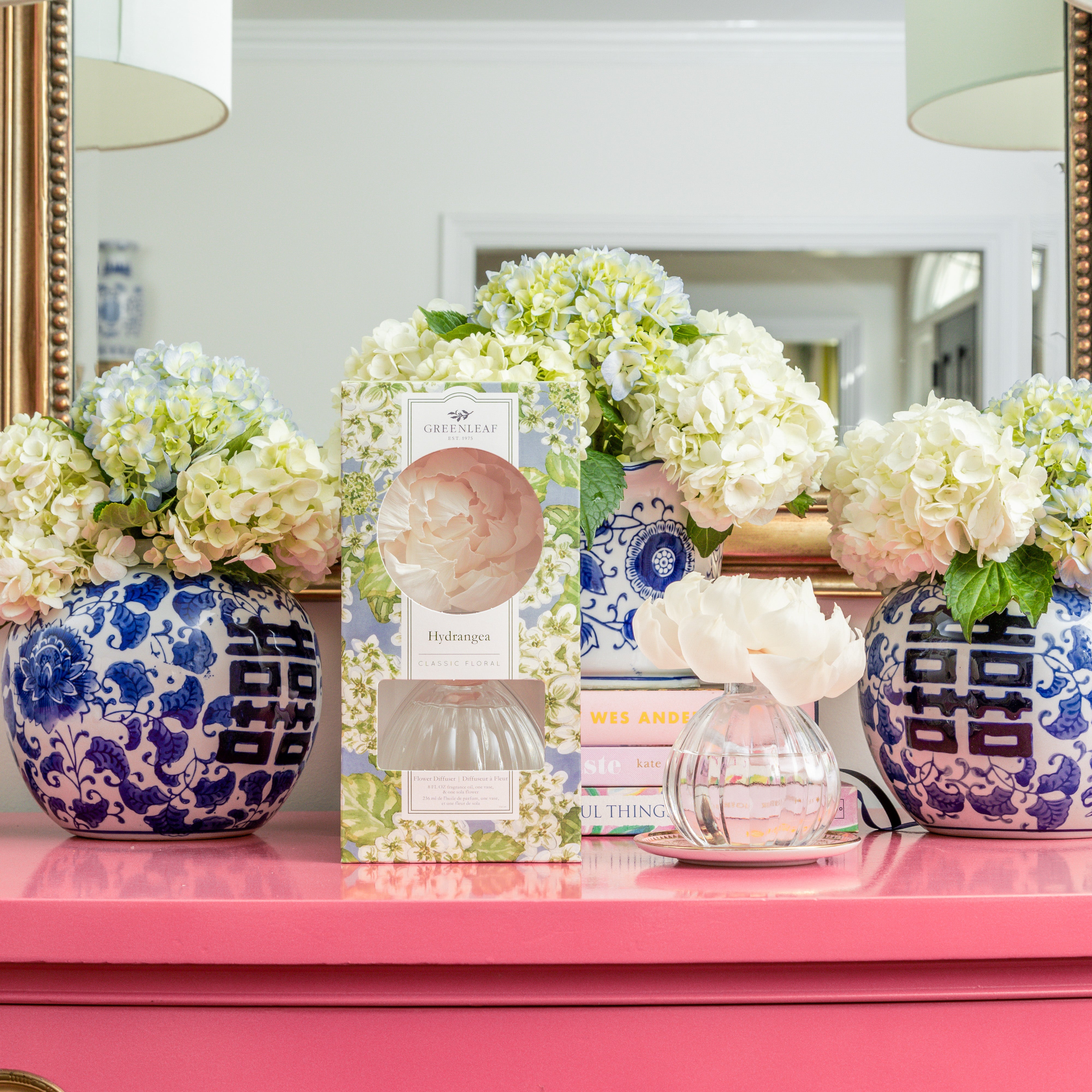 A pink table holds Greenleaf Gifts' Hydrangea Flower Diffuser, featuring sola flowers and hydrangea-scented oil in a flower-shaped bottle, plus blue and white vases with white hydrangeas—all arranged before a mirror.