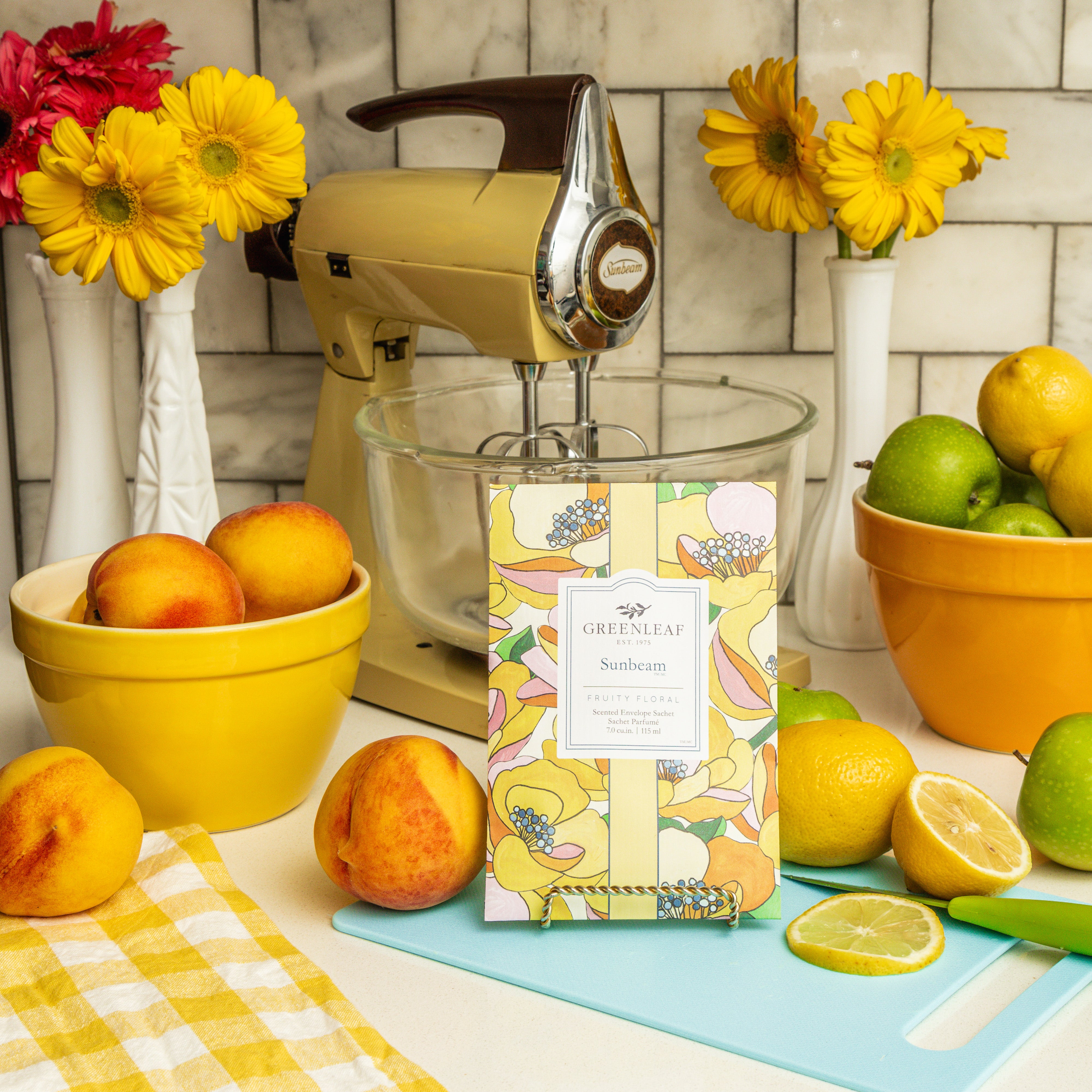 A bright kitchen scene with yellow flowers, a stand mixer, peaches, lemons, and limes. The Greenleaf Gifts Sunbeam Sachet sits in front on a yellow checkered cloth with fruit—an air freshener that brings long-lasting fragrance.