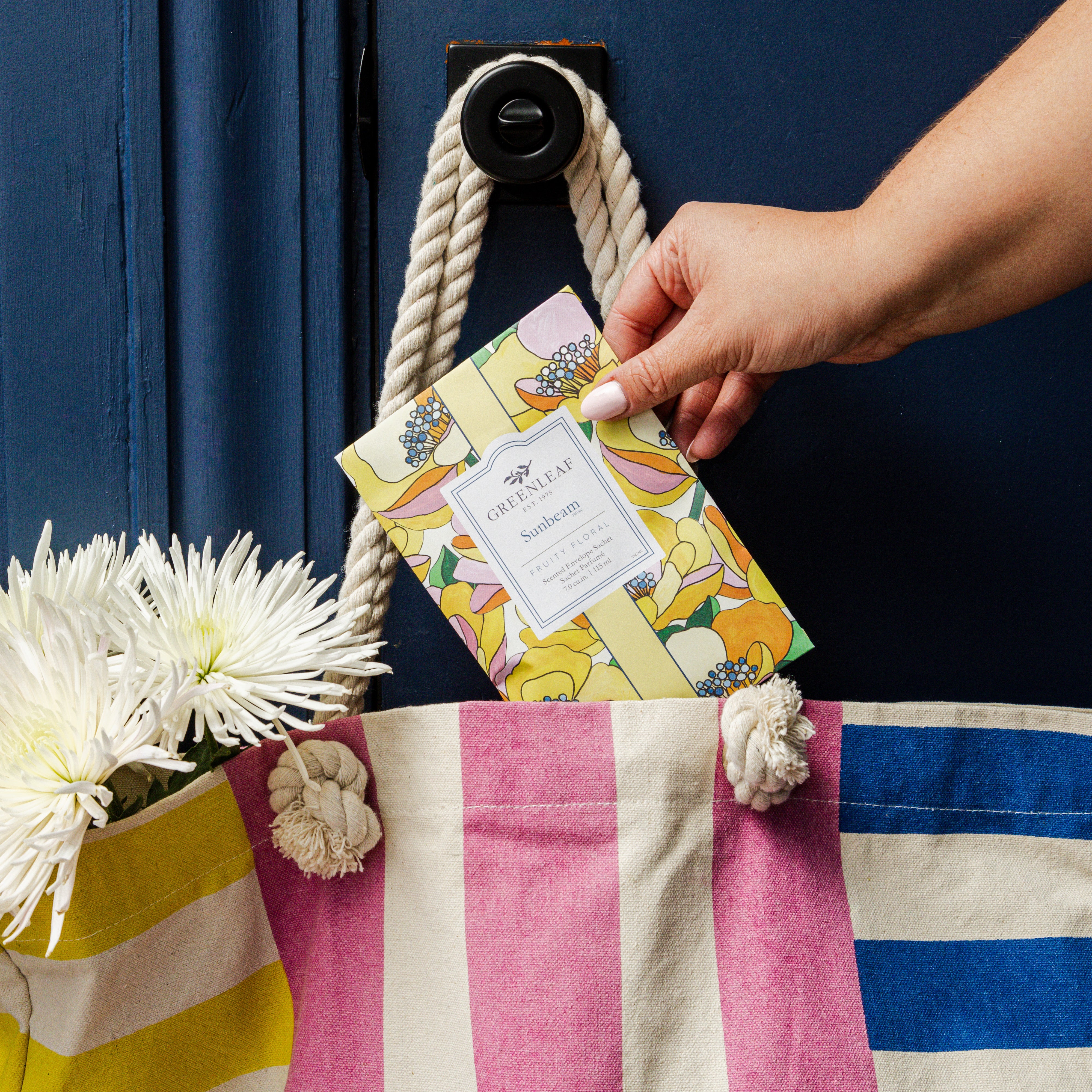 A hand places a Greenleaf Gifts Sunbeam Sachet into a striped tote with pink, yellow, and white sections on a dark blue door. The bag, holding white flowers, gains long-lasting freshness from the uplifting Sunbeam fragrance.