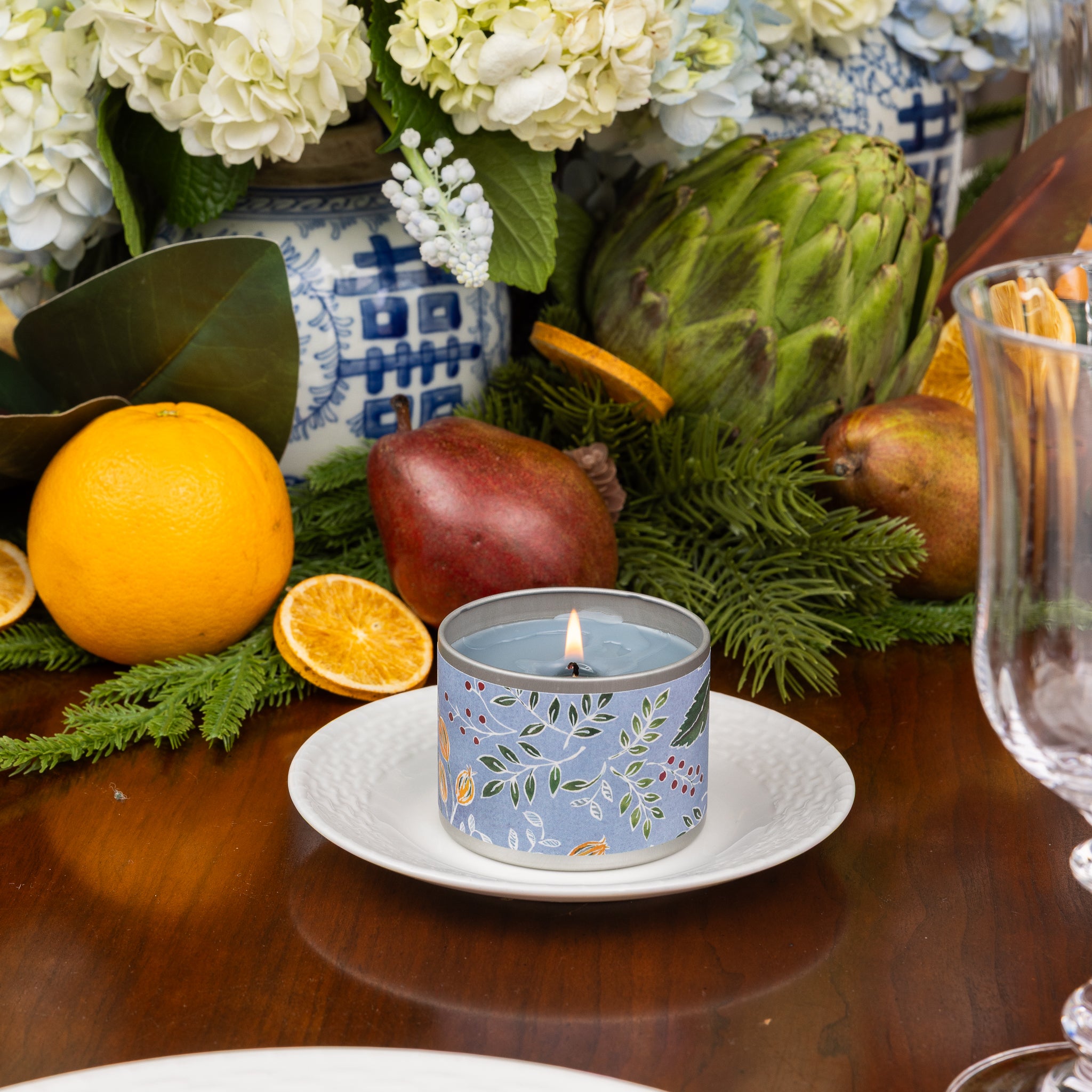 A lit Laurel Grove Tin Candle by Greenleaf Gifts glows in its decorative blue tin atop a white plate, surrounded by fresh oranges, pomegranate, artichoke, greenery, and blue and white vases filled with flowers on a wooden table. This delightful arrangement brings the inviting aroma of Greenleaf Gifts to your space—perfect for brightening any room.