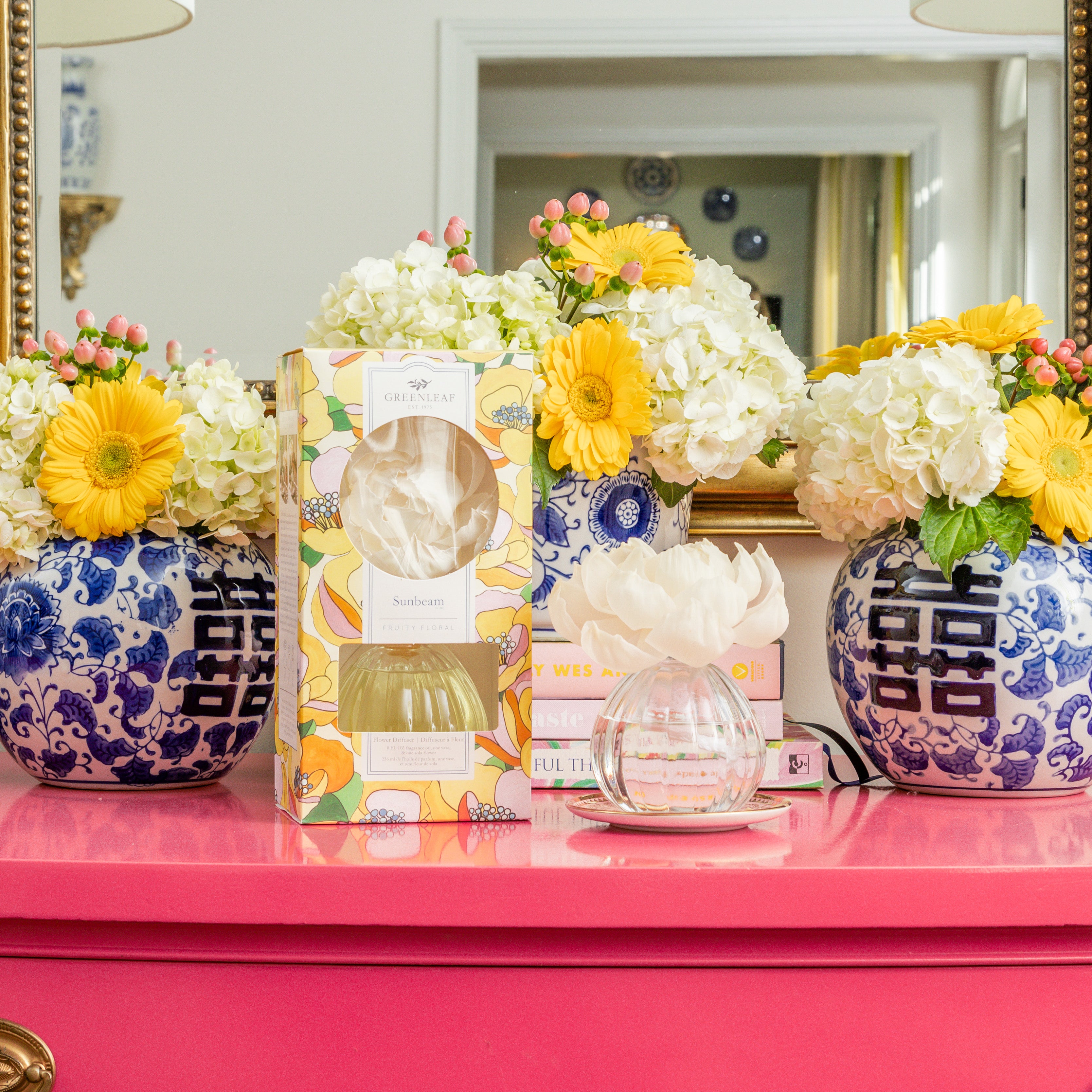 A bright pink table displays two blue and white vases with yellow flowers and hydrangeas, a floral box, a stack of books, and the Greenleaf Gifts Sunbeam Flower Diffuser with fragrance oil; a mirror reflects the scene behind them.