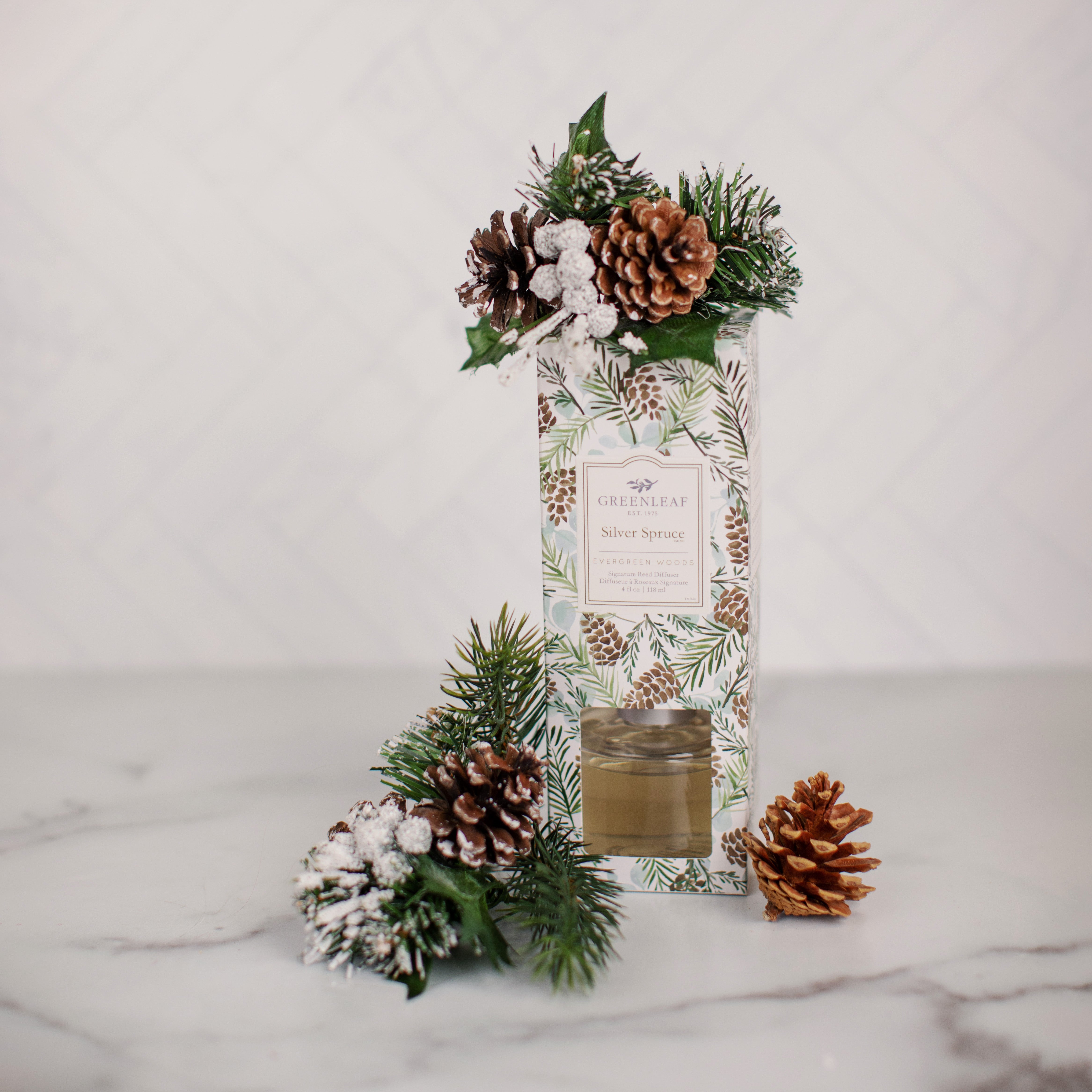The Greenleaf Gifts Silver Spruce Reed Diffuser features a festive box with pine and leaf motifs, frosted accents, pinecones, and greenery. Displayed on marble with a pinecone, it offers the refreshing scent of Silver Spruce.