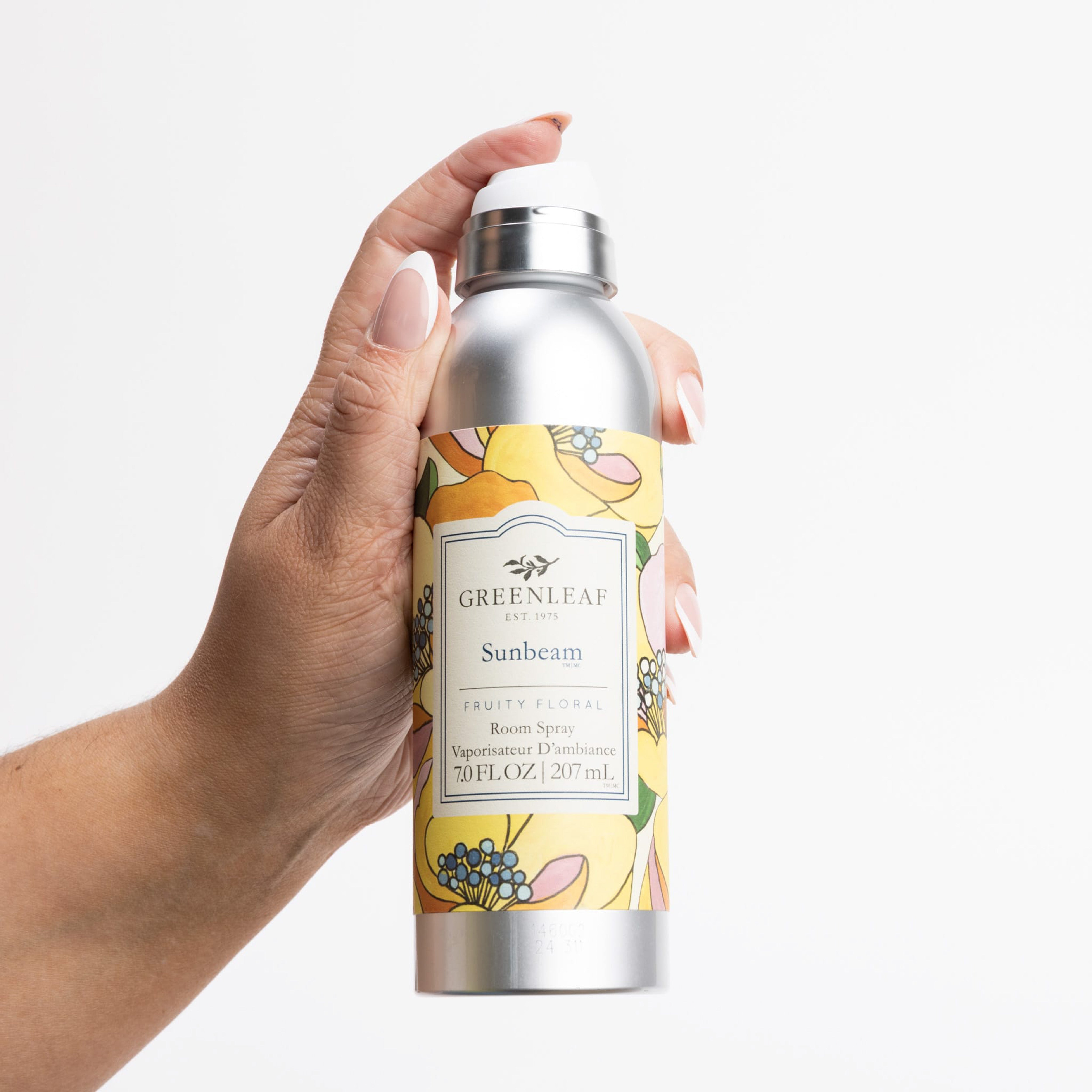 A hand with light pink nail polish holds a 7 fl oz (207 ml) Sunbeam Room Spray by Greenleaf Gifts, featuring a yellow floral design and labeled as a fruity floral fragrance.