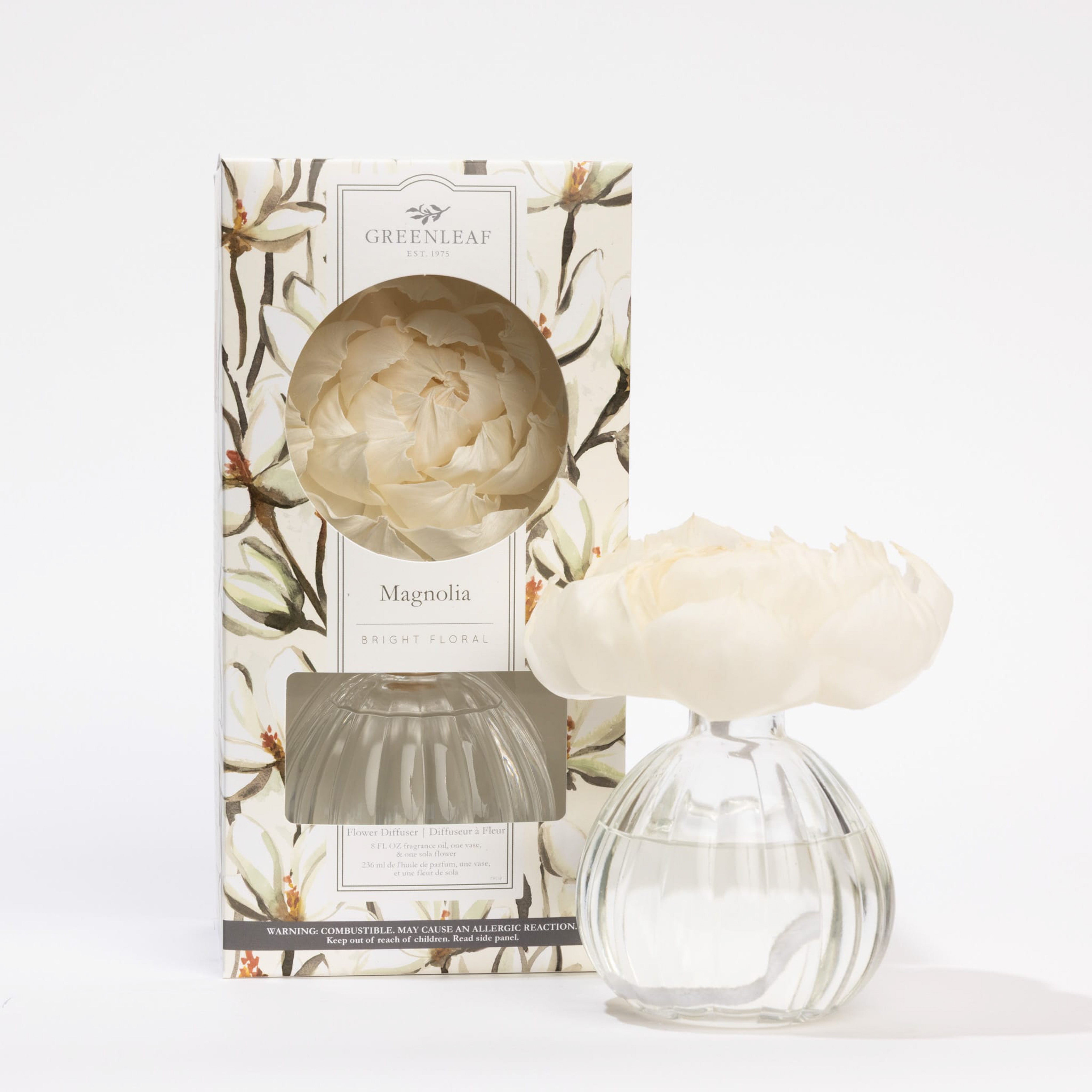 The Greenleaf Gifts Flower Diffuser-Magnolia, a glass vase with a white magnolia flower, provides 60 days of fragrance and sits beside its 8 oz floral-patterned box (GLG966515S) on a white background.