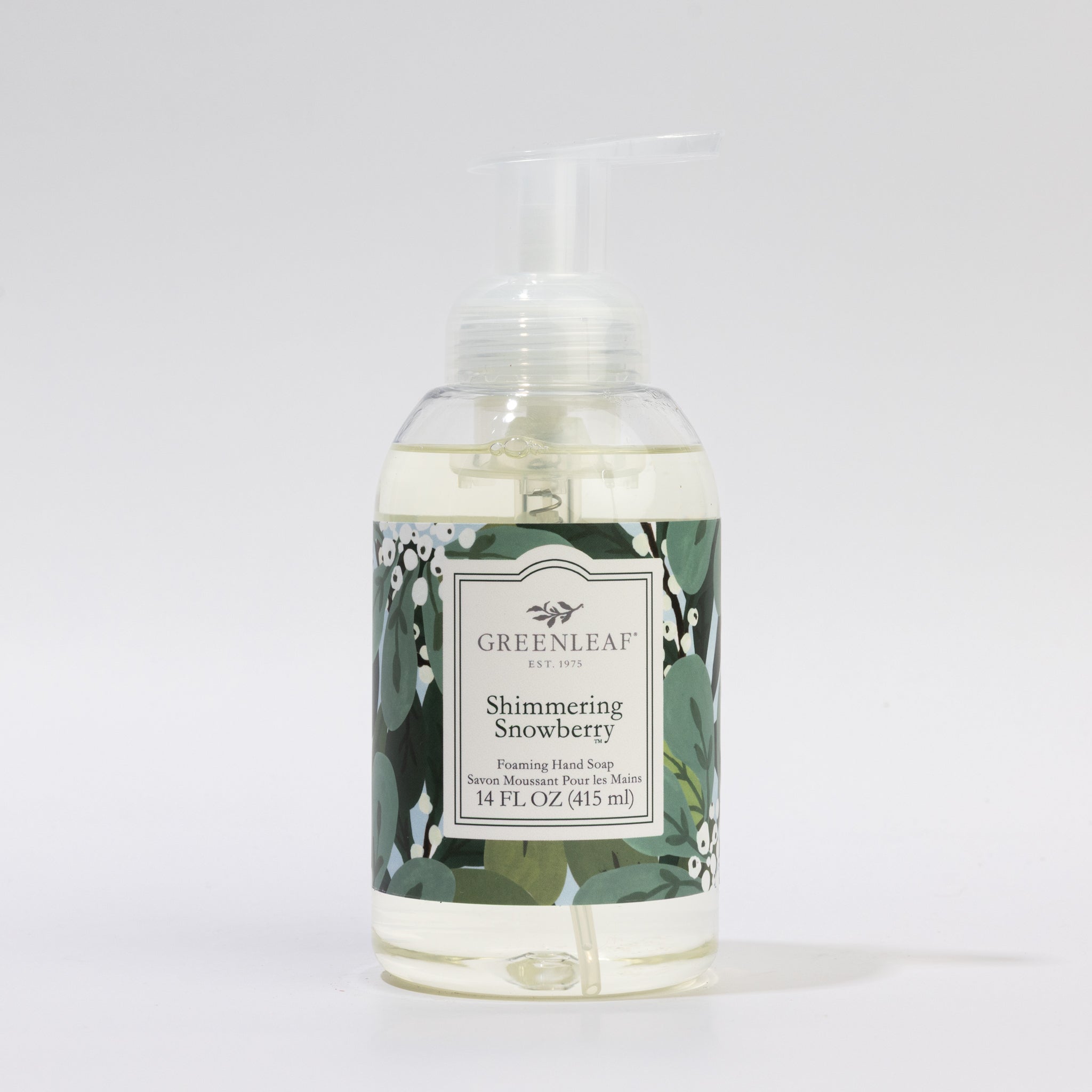 A clear pump bottle of Greenleaf Gifts Shimmering Snowberry Foaming Hand Soap, 14 fl oz (415 ml), features green leaves and white berries on a dark label—an elegant addition beside your favorite 14 oz glass.