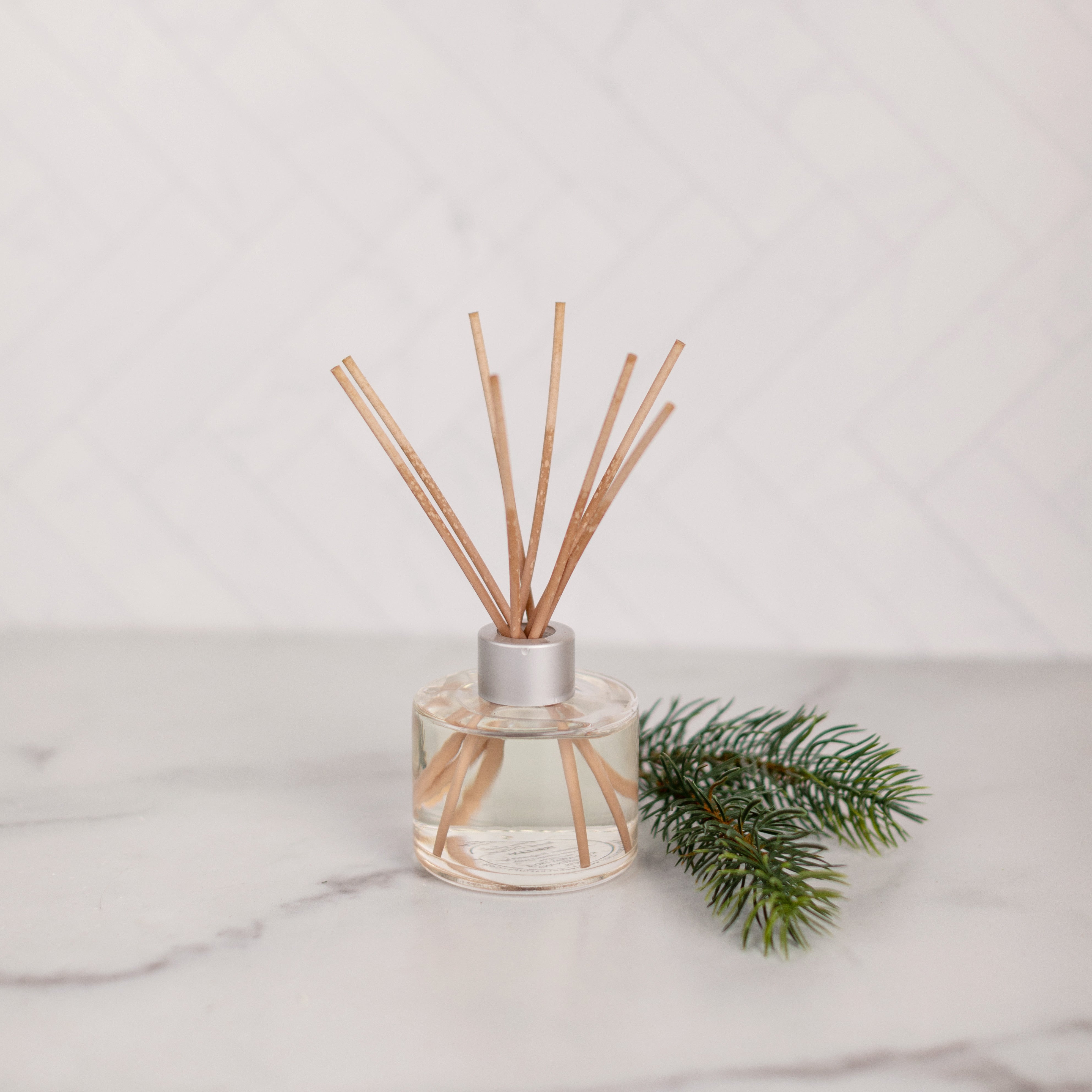 The Greenleaf Gifts Silver Spruce Reed Diffuser, featuring light fiber reeds, rests on a white marble surface beside a green pine branch, filling the space with its fresh aroma against a light, textured background.