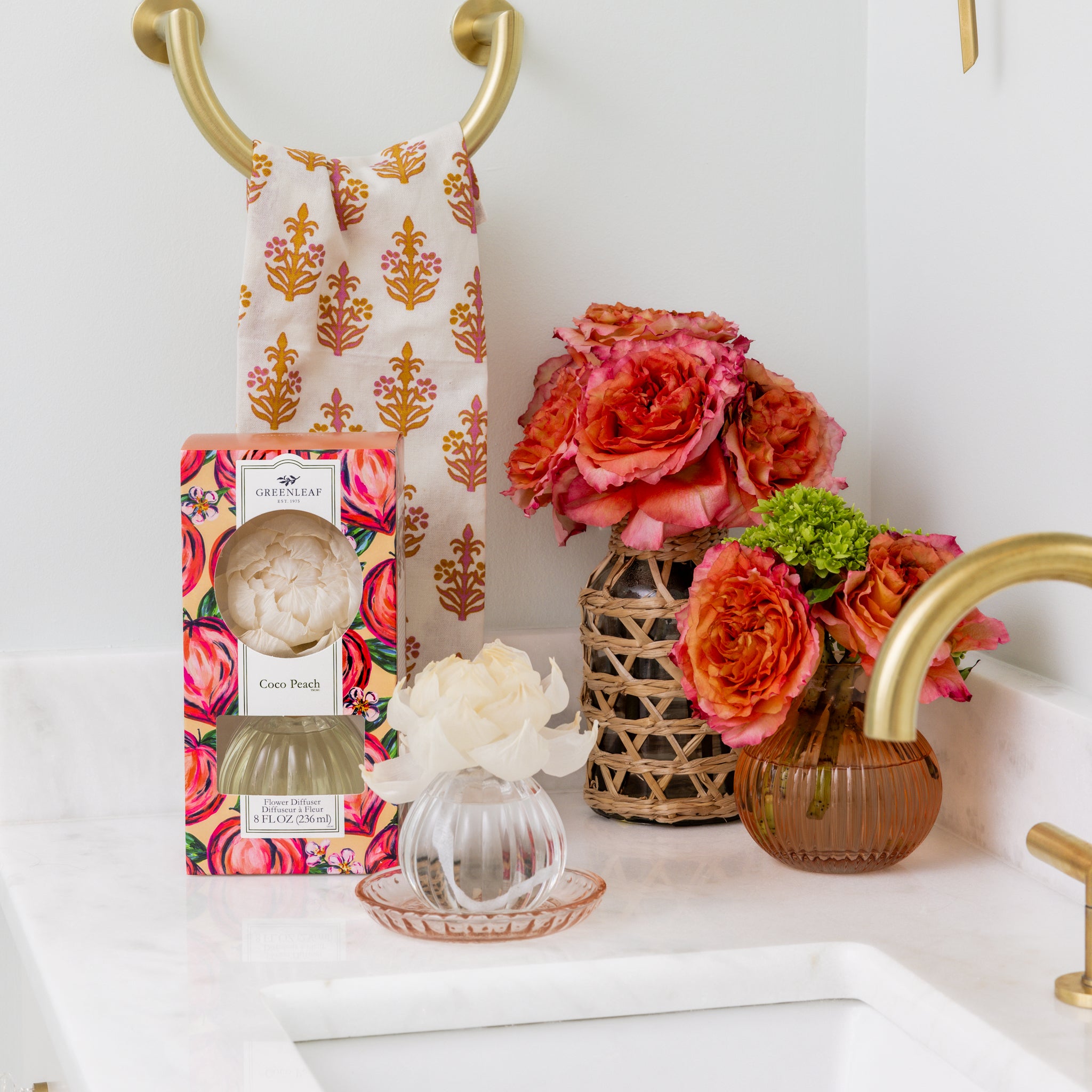 A bathroom countertop with gold fixtures, a floral towel on a ring, flower-shaped soap, the Greenleaf Gifts Coco Peach Flower Diffuser, and two vases of bright pink and orange flowers.
