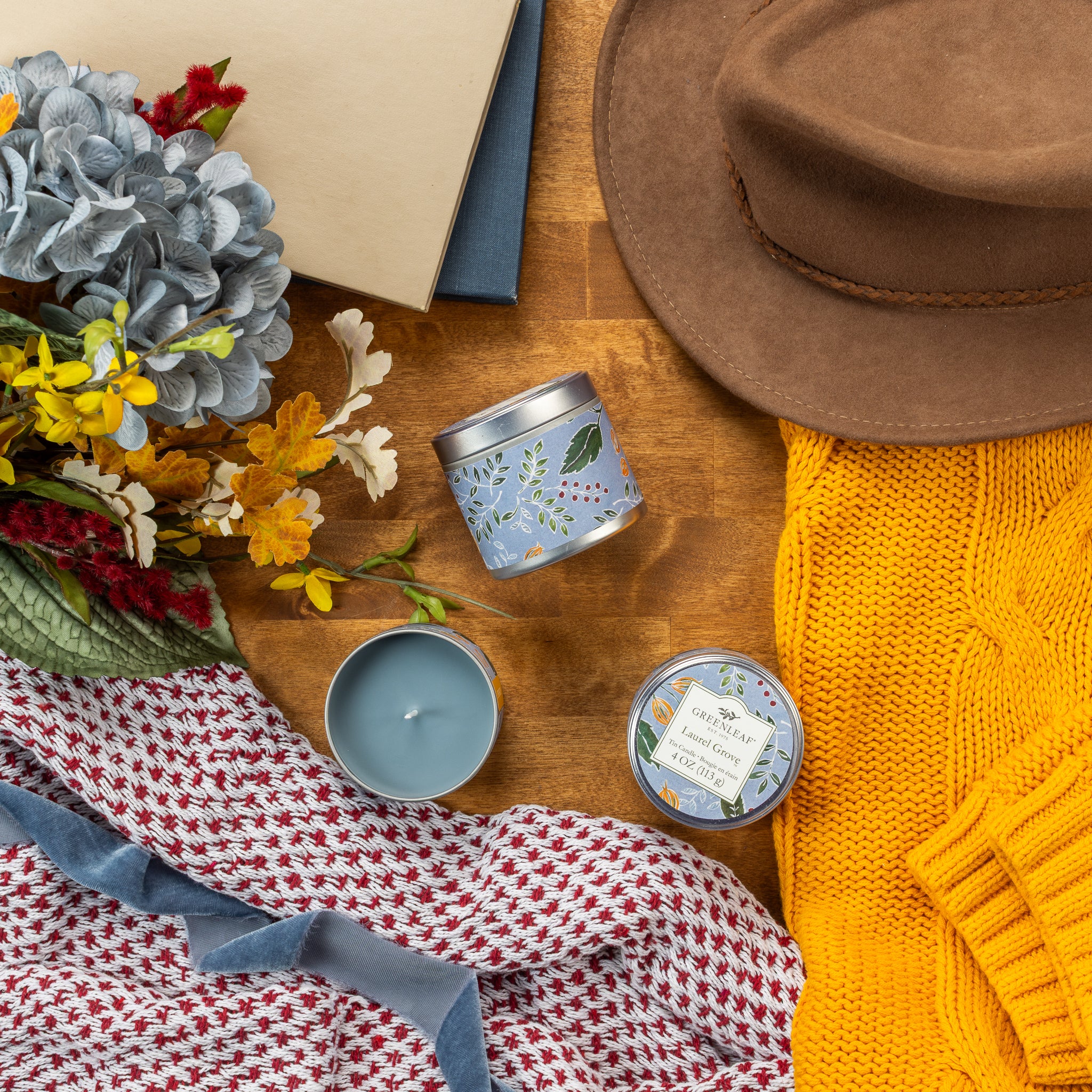A cozy flat lay with a brown hat, yellow sweater, two Greenleaf Gifts Laurel Grove Tin Candles, an open book, a bouquet of flowers, and a patterned red and white scarf on a wooden surface.