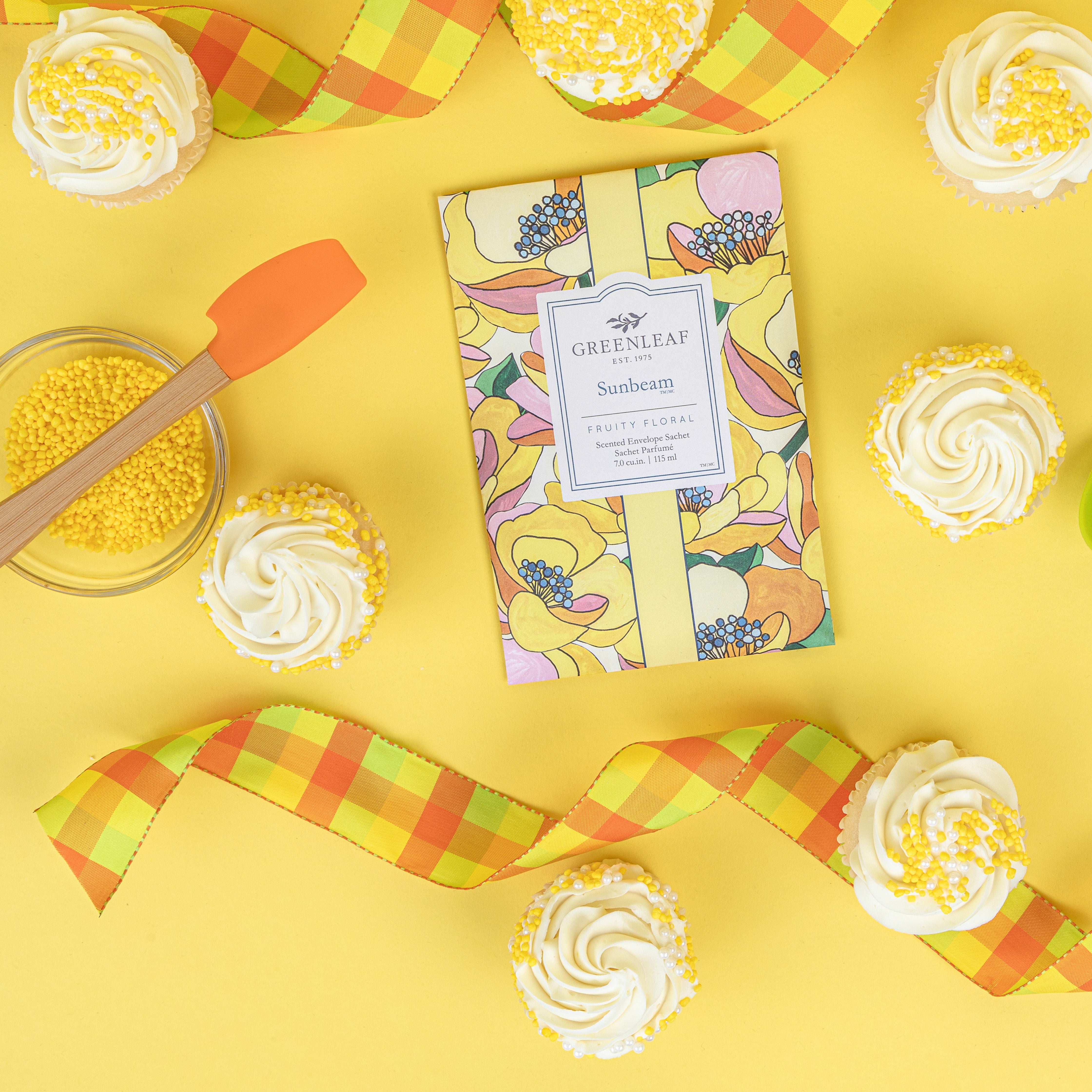 A vibrant Greenleaf Gifts Sunbeam Sachet is surrounded by white-frosted cupcakes with yellow sprinkles, a bowl of yellow sprinkles with a spatula, and plaid yellow-orange ribbons on a bright yellow background.