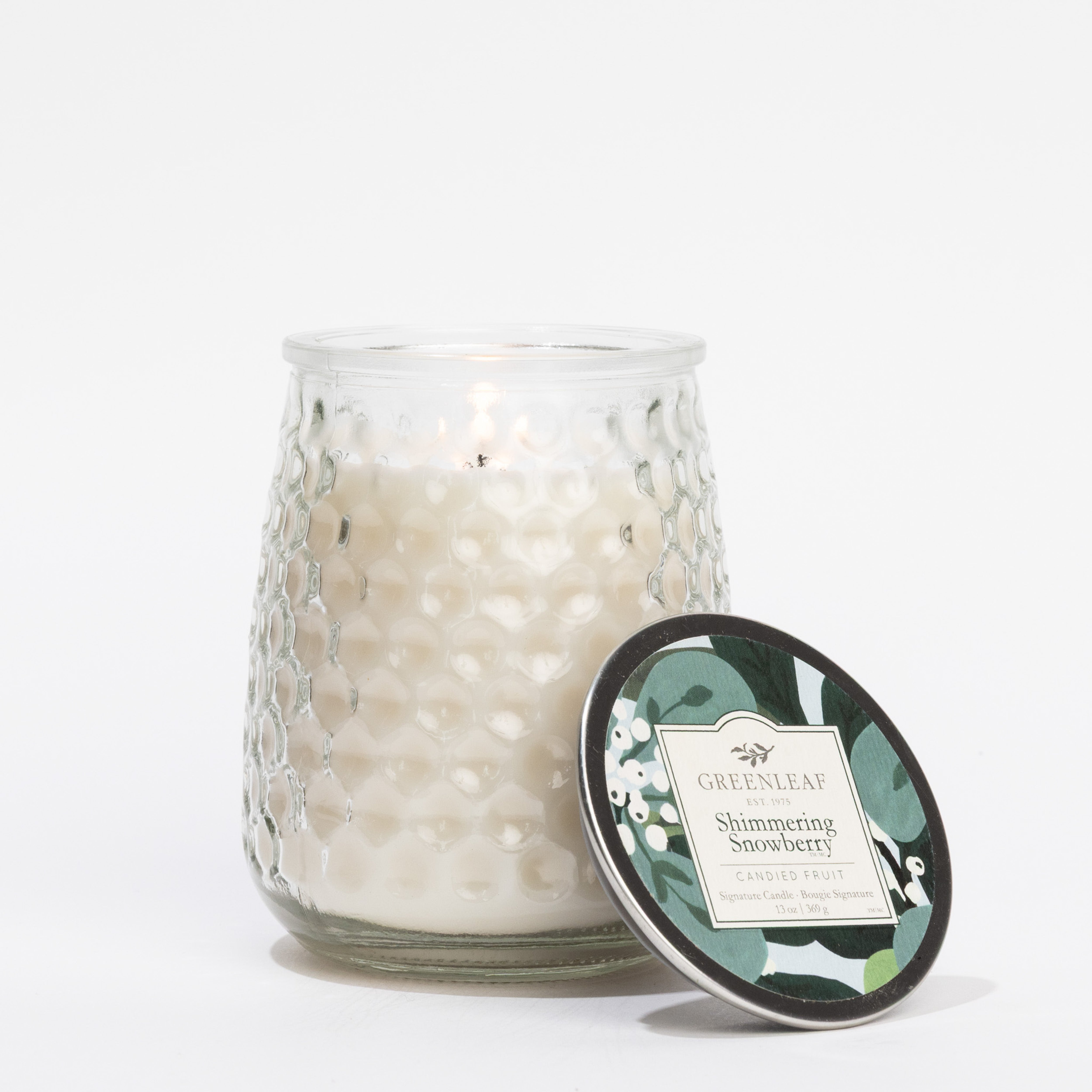 A lit Shimmering Snowberry Signature Candle by Greenleaf Gifts in a textured glass jar sits beside its lid, featuring a green and white botanical label—an inviting touch from Greenleaf Gifts fragrances.