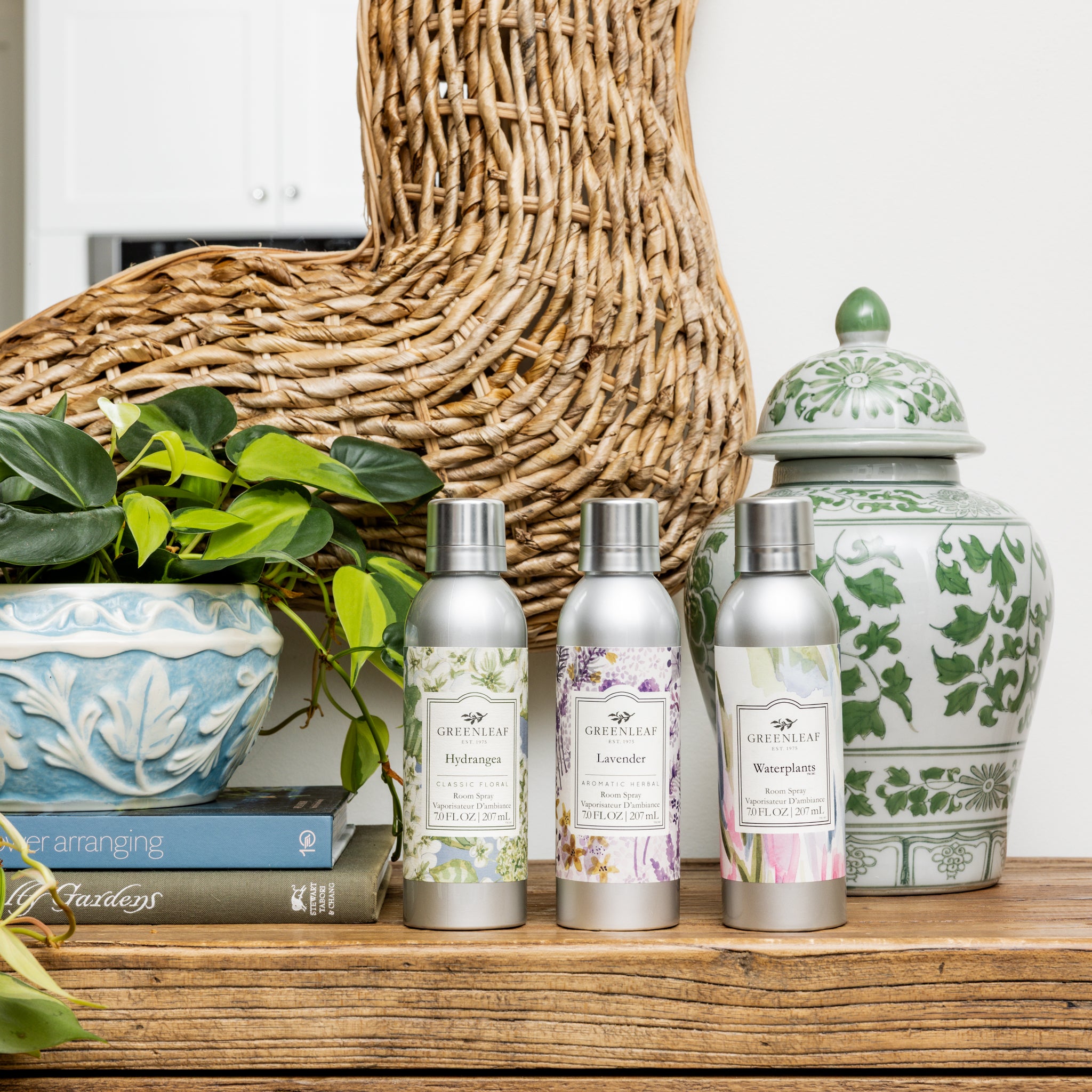 Three Greenleaf Gifts Waterplants Room Spray bottles sit on a wooden table with decorative items, such as a leafy potted plant, green and white ceramics, a woven basket, and stacked books in the background.