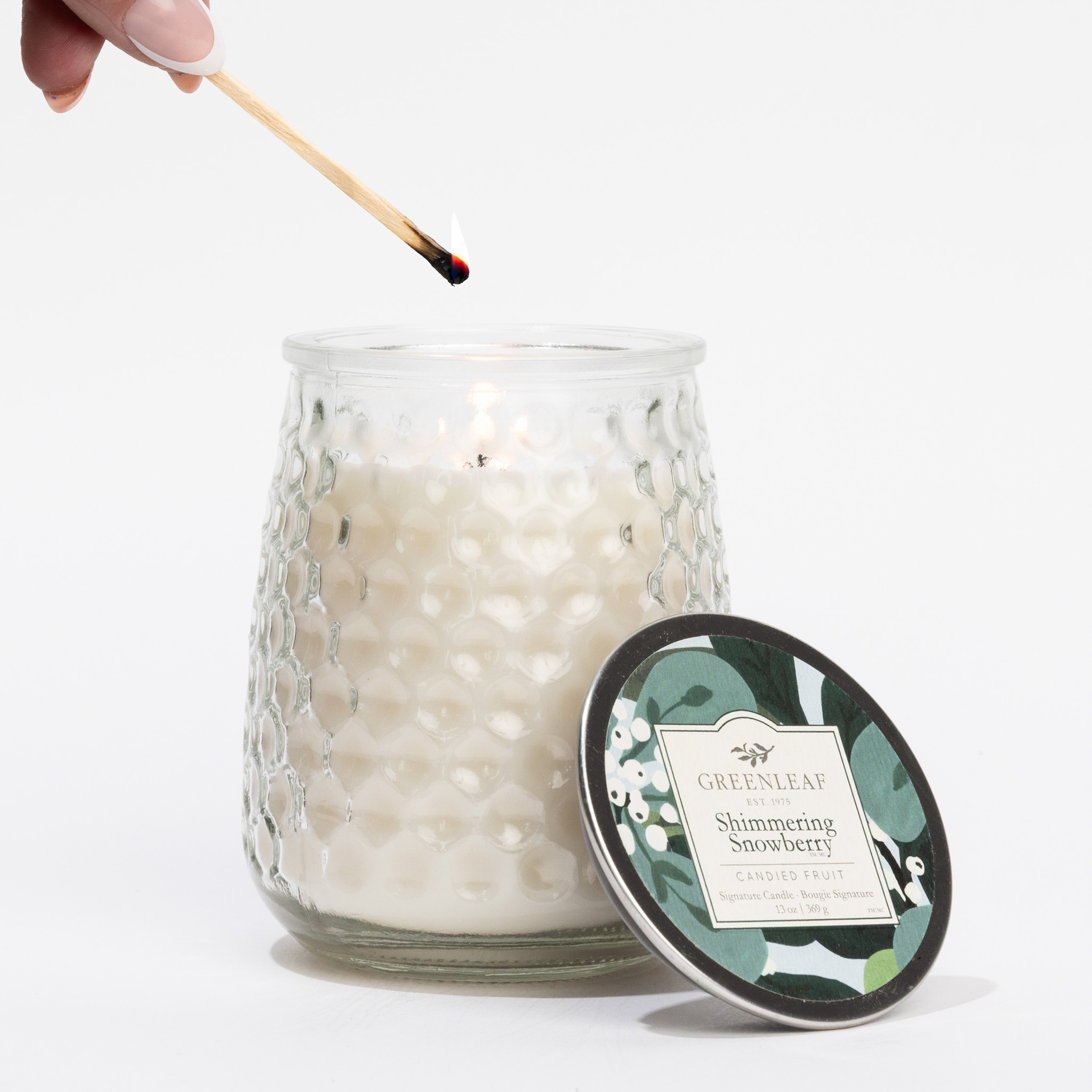 A hand lights a white Shimmering Snowberry Signature Candle by Greenleaf Gifts, housed in a textured glass jar. The candle’s green-and-white leaf lid rests nearby.