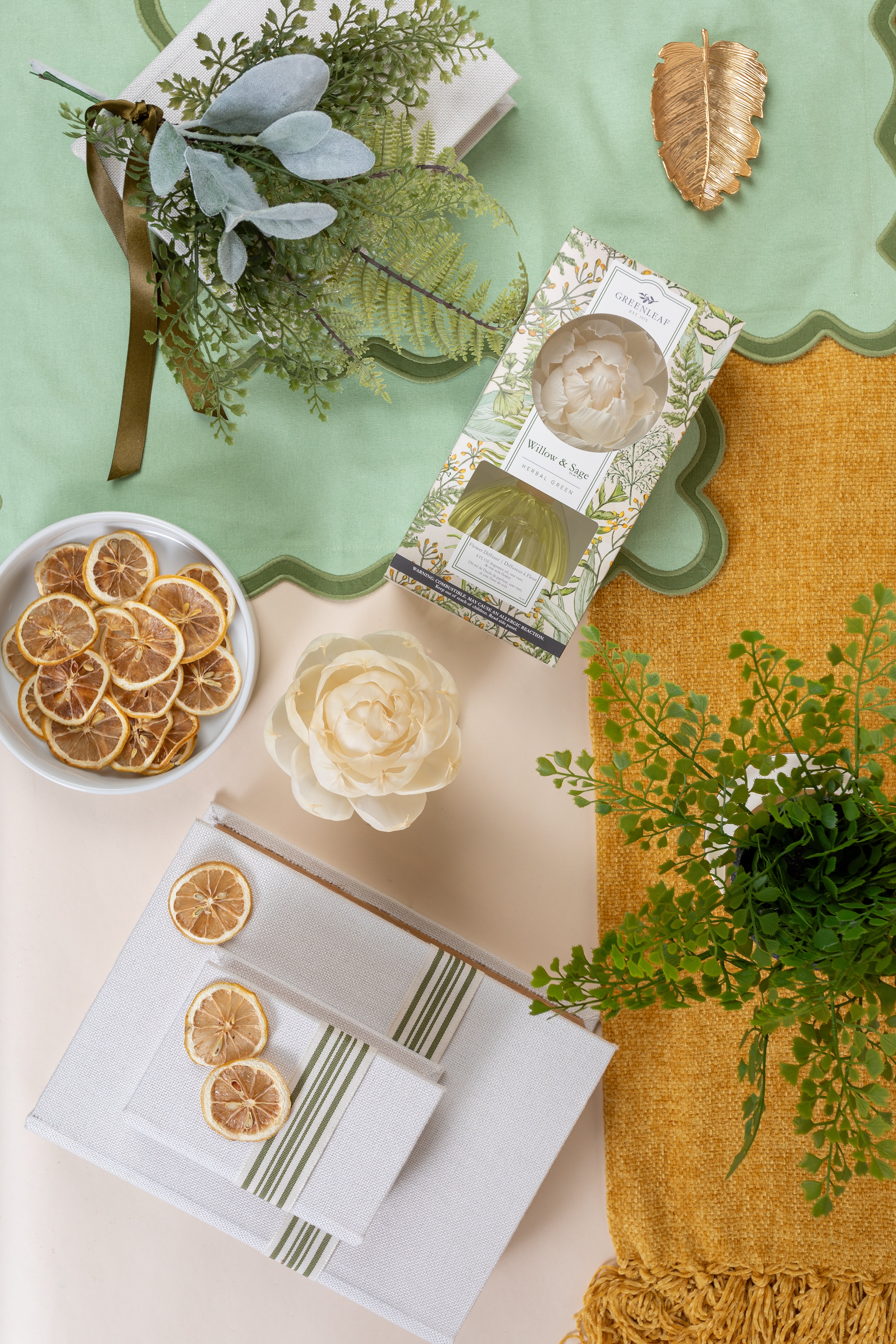 A flat lay of a table set with a green and yellow scalloped tablecloth, Greenleaf Gifts' Willow & Sage Flower Diffuser (8 oz), a rose-shaped white accent, dried orange slices, green foliage, gold leaf ornament, and folded napkins.