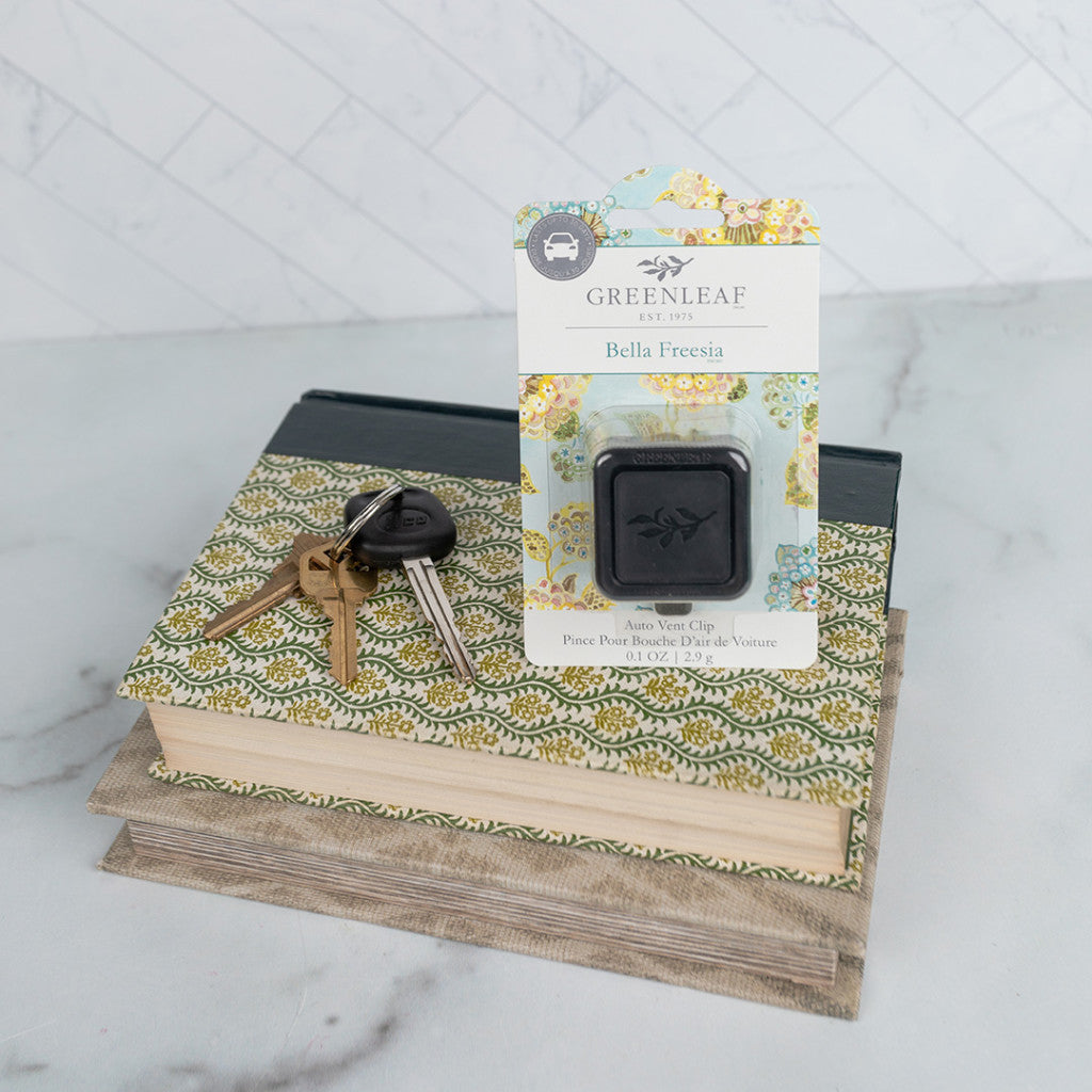 A set of keys and a Greenleaf Gifts Bella Freesia Auto Vent Clip rest on two patterned decorative books against a light, tiled background.