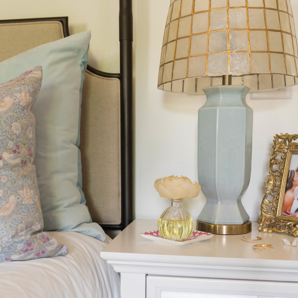 A bedside table holds a large blue lamp, a framed photo, jewelry, and a Greenleaf Gifts Spa Springs Flower Diffuser. The bed features pale blue pillows and a decorative floral-patterned cushion.