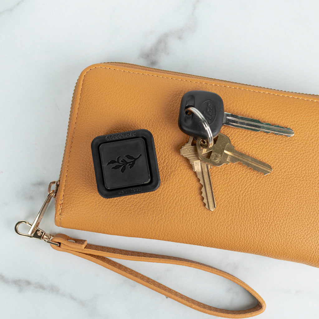 A tan leather wristlet with a zipper and strap rests on a white surface. On top are a black key fob with a leaf design, three keys on a black keychain, and the Greenleaf Gifts Bella Freesia Auto Vent Clip.