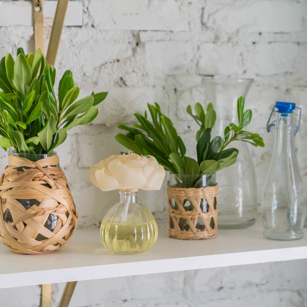 A white shelf displays glass and woven vases with green leafy plants, a pale flower, and Greenleaf Gifts Spa Springs Flower Diffuser bottles, all set against a crisp white brick wall.